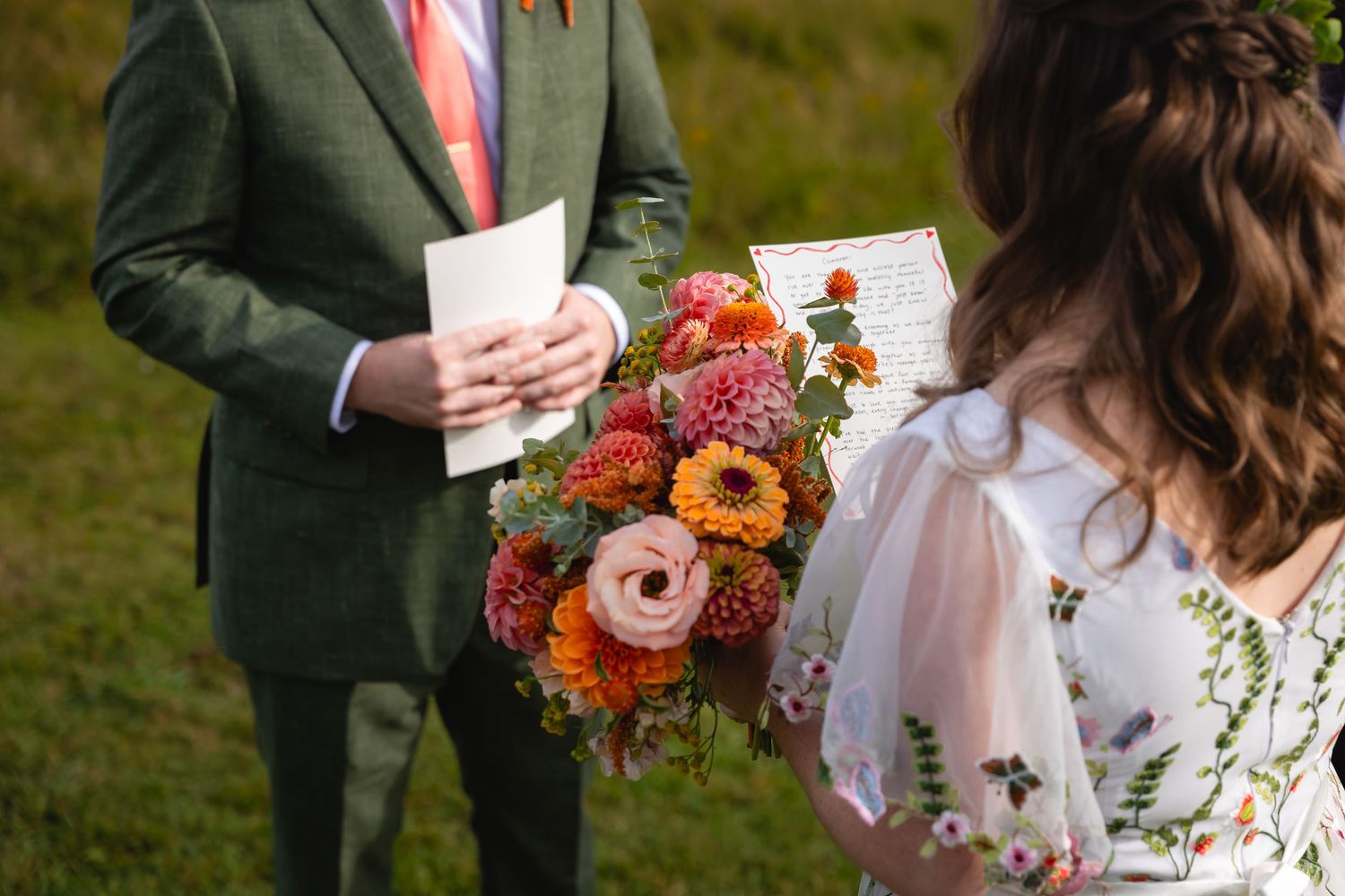 A wedding couple stands together outdoors holding vibrant autumn-colored bouquets with orange and pink flowers.