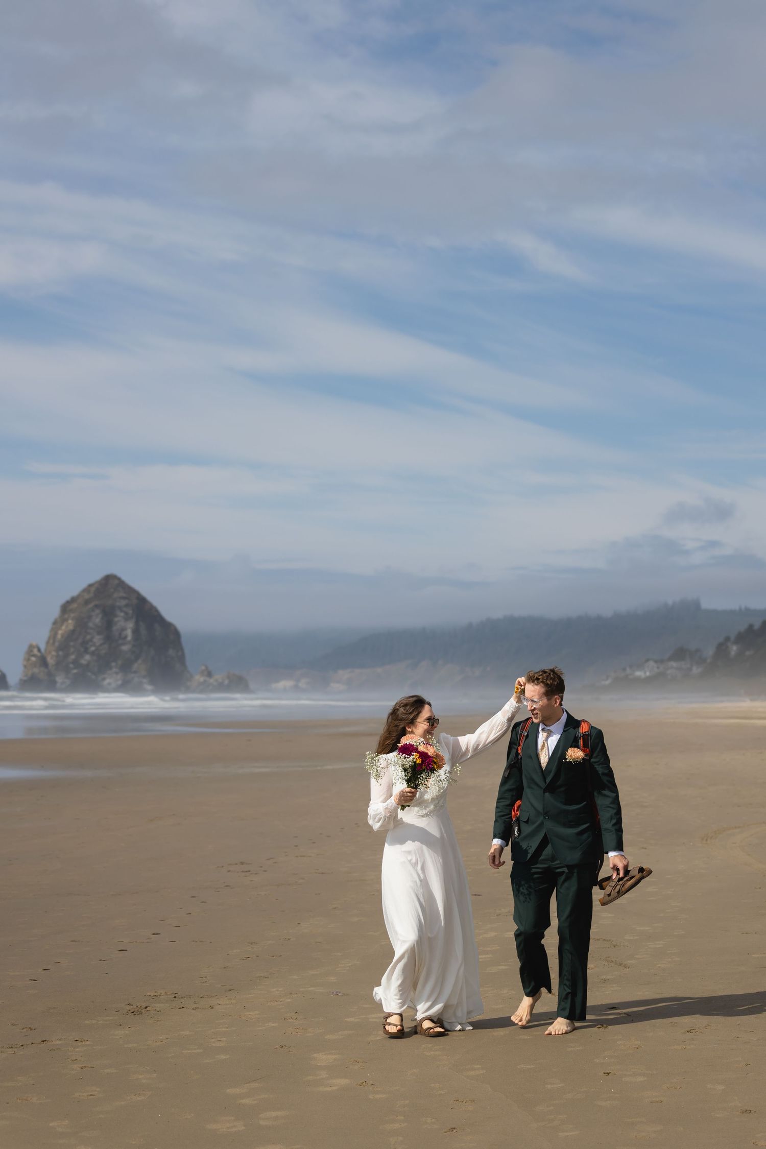 A couple walks along a misty beach with dramatic coastal rock formations visible in the distance on a cloudy day.