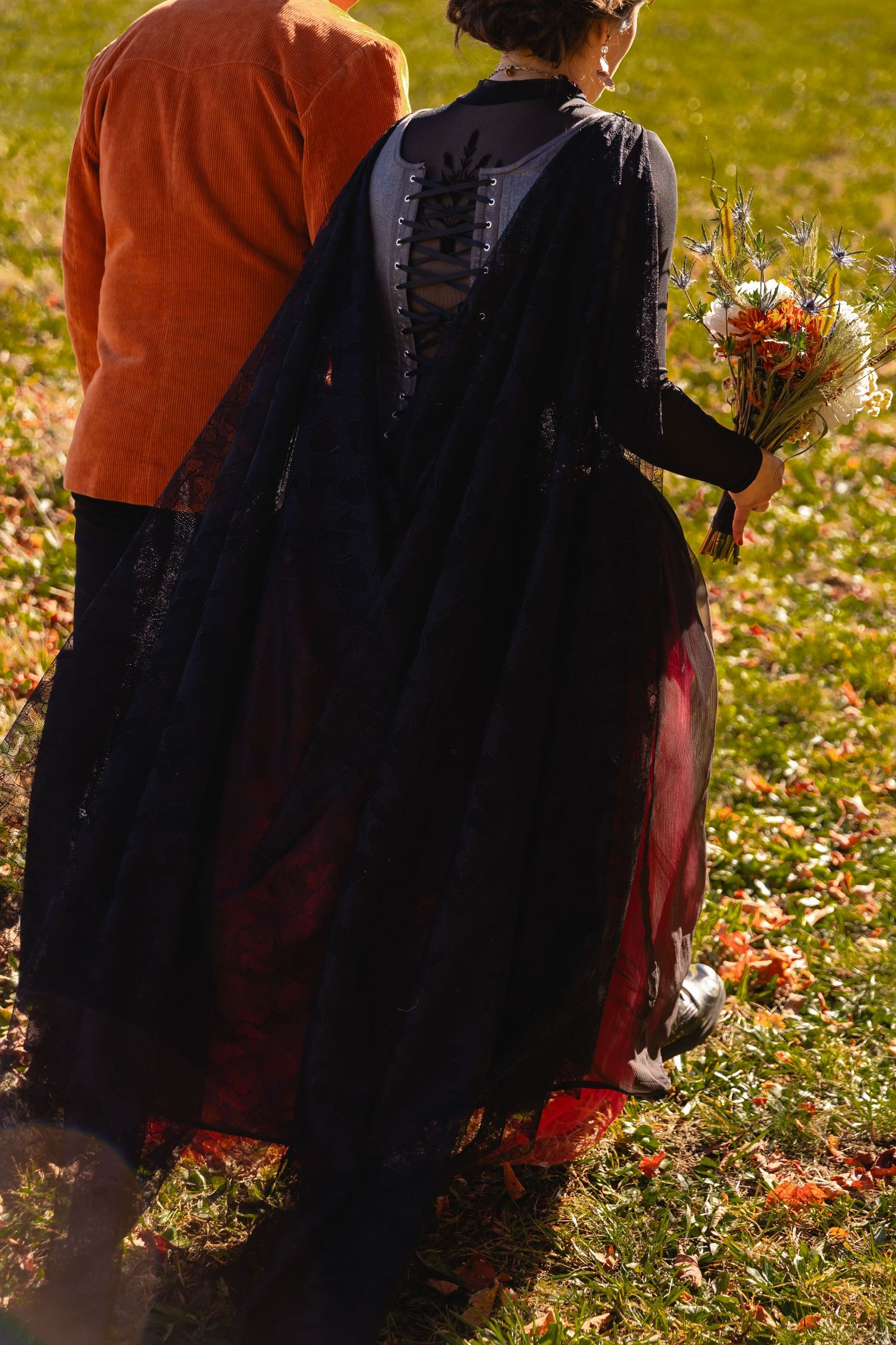 Two figures in contrasting orange and black clothing walk together through autumn leaves in a sunlit outdoor setting.