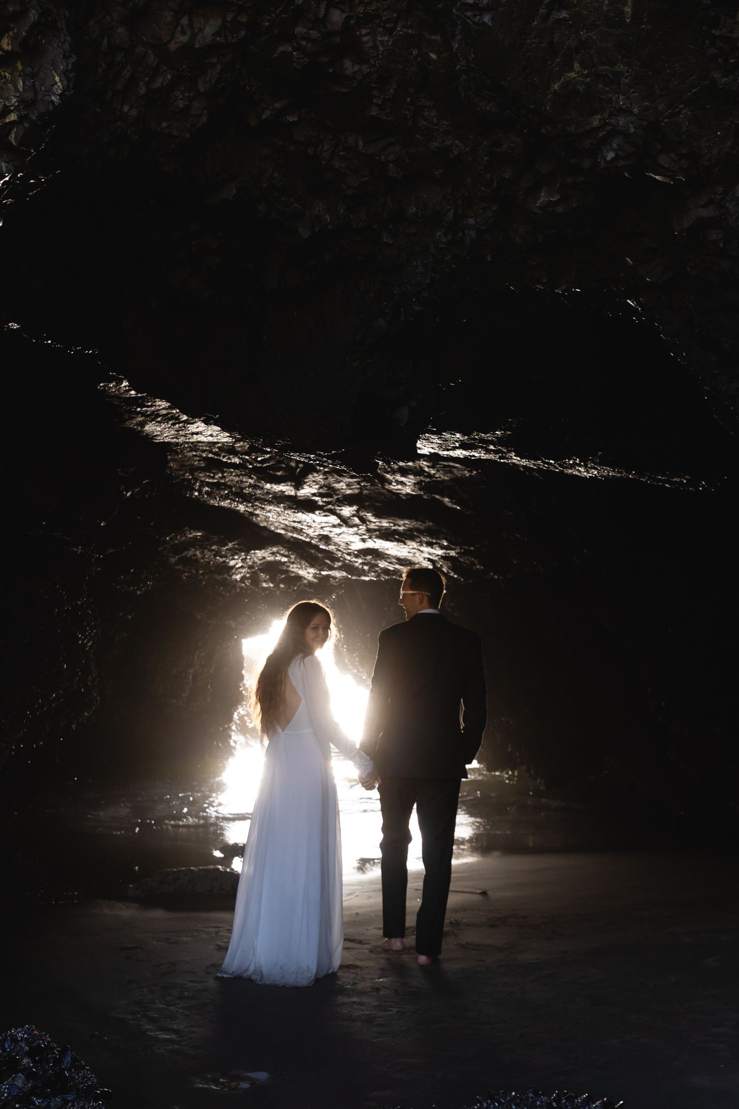 A couple in wedding attire is silhouetted against dramatic backlighting in a dark cave setting.