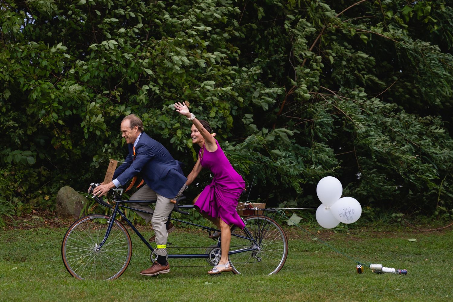 Couple riding tandem bicycle on grass, woman in purple dress waving joyfully.