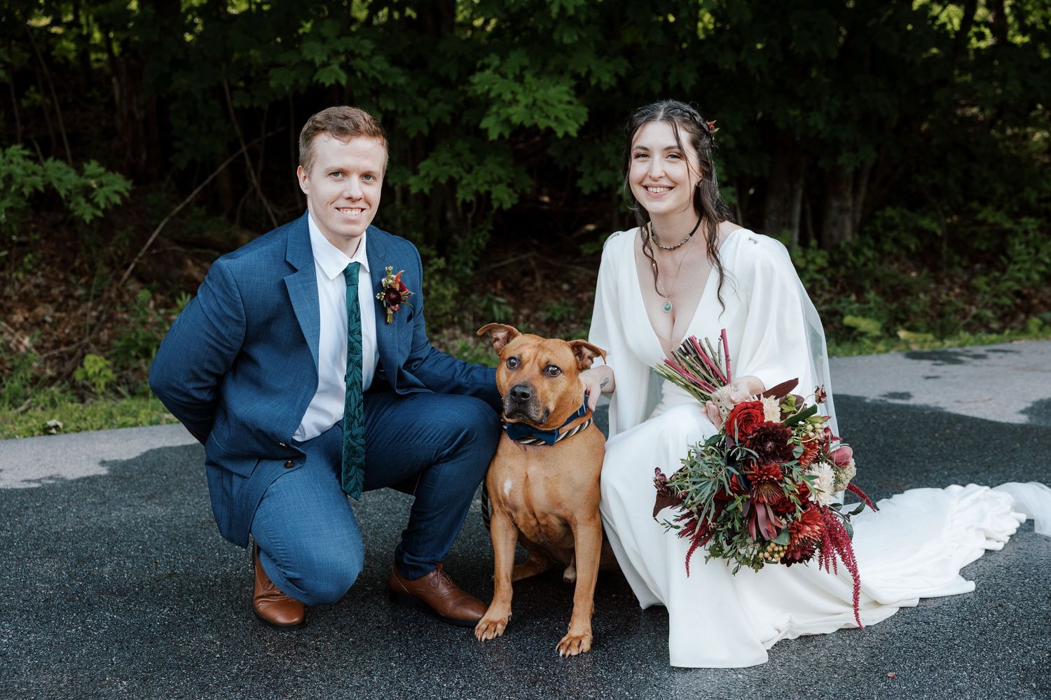 A bride and groom kneel beside their brown dog for a wedding portrait outdoors.