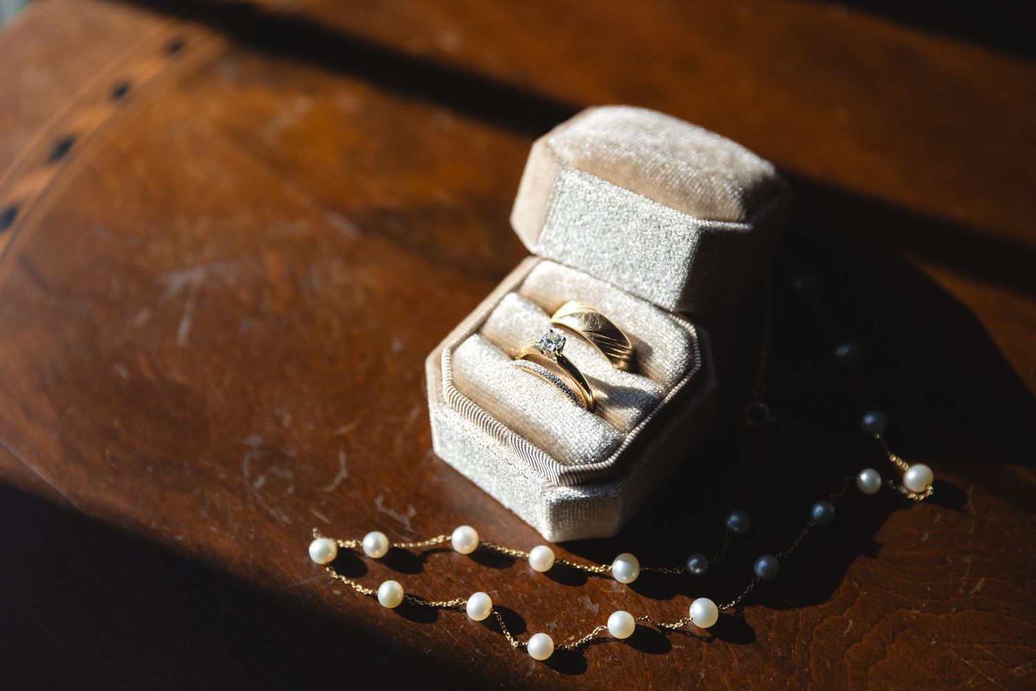 Wedding rings nestled in a white box alongside delicate pearl jewelry on a dark wooden surface.
