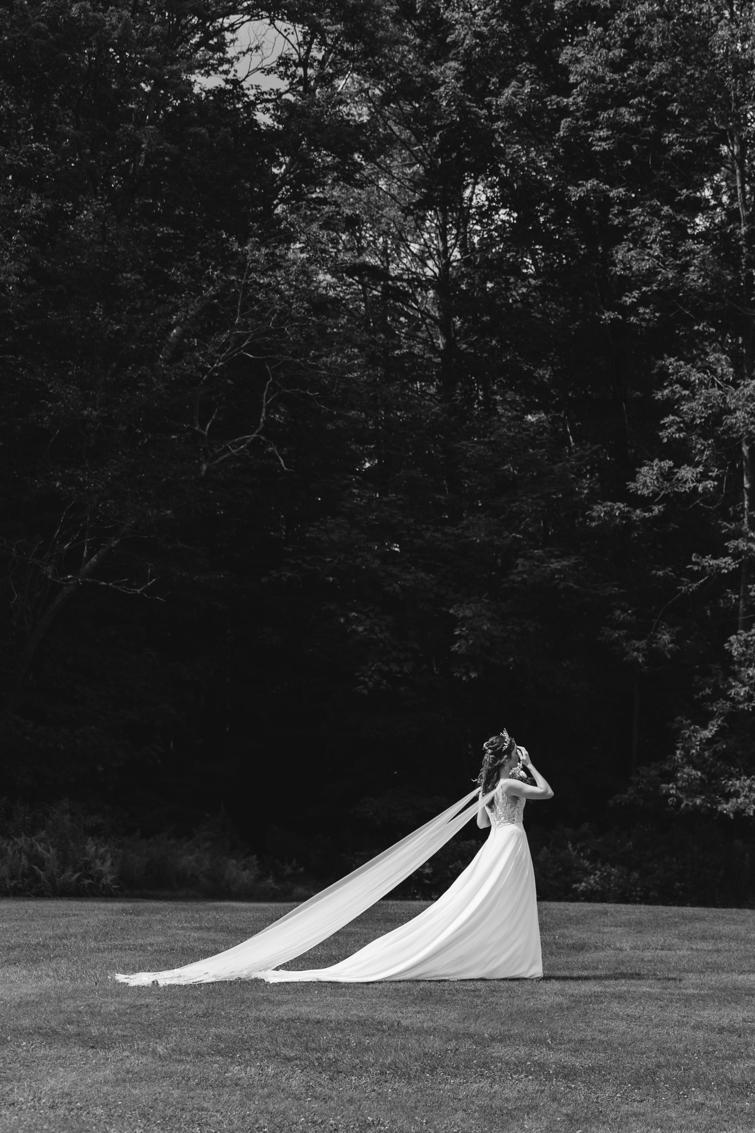 A dramatic black and white photo of a bride's flowing dress train on a grassy field against dark woods.