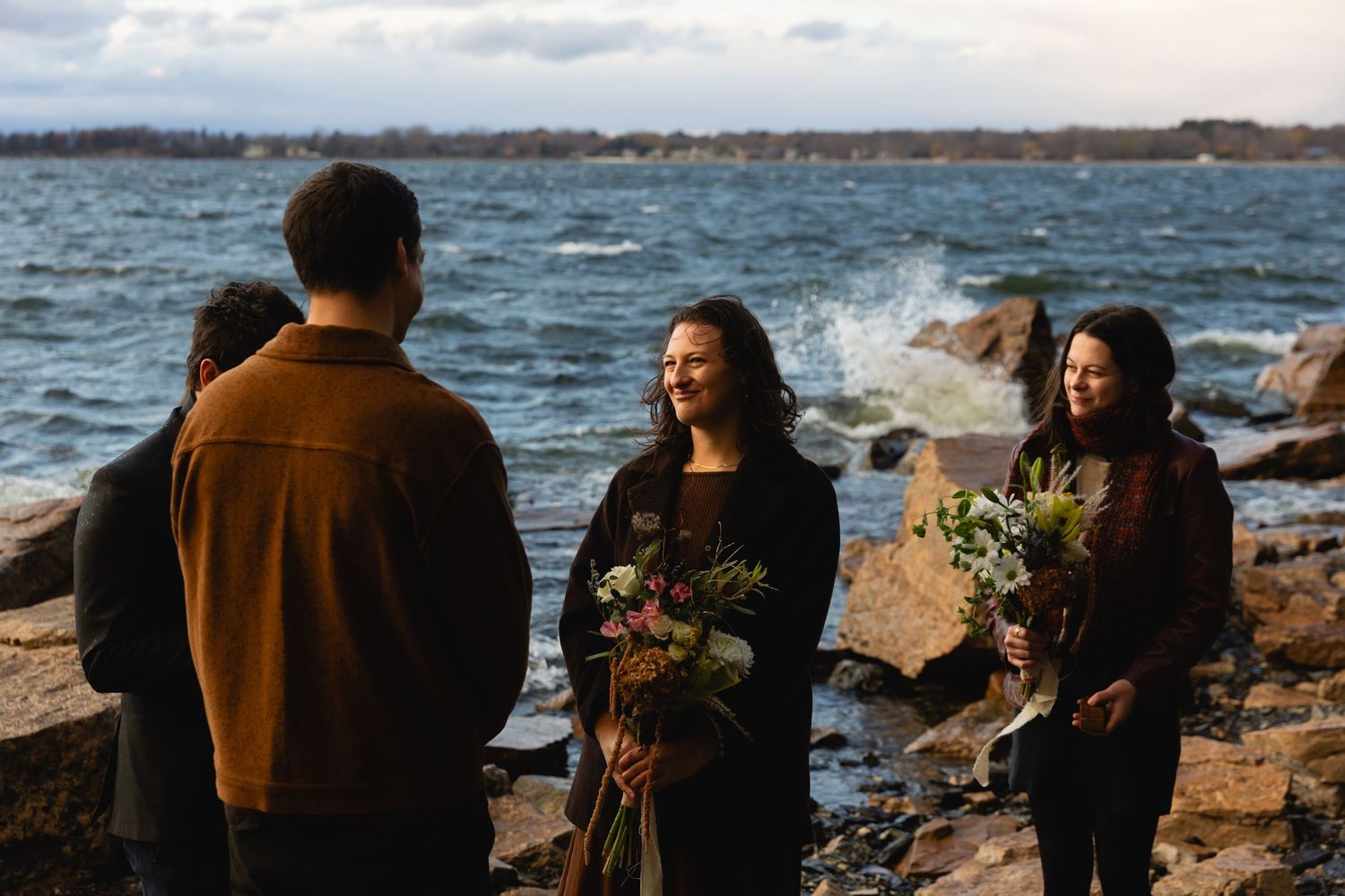 A small group participates in a seaside gathering among rocky outcrops with bouquets of wildflowers.
