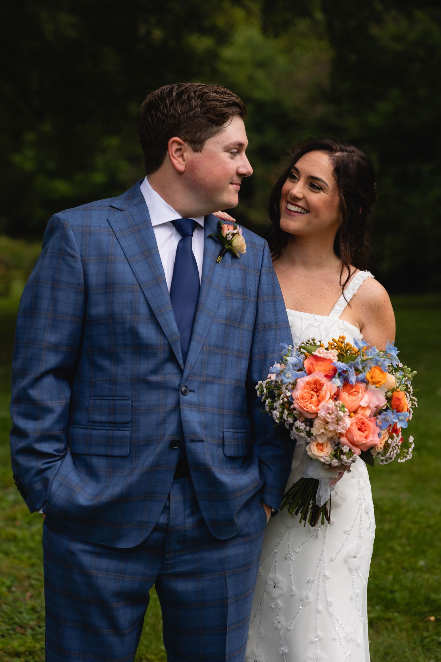 A couple enjoys a joyful moment together outdoors, with the bride holding a vibrant coral and blue bouquet.