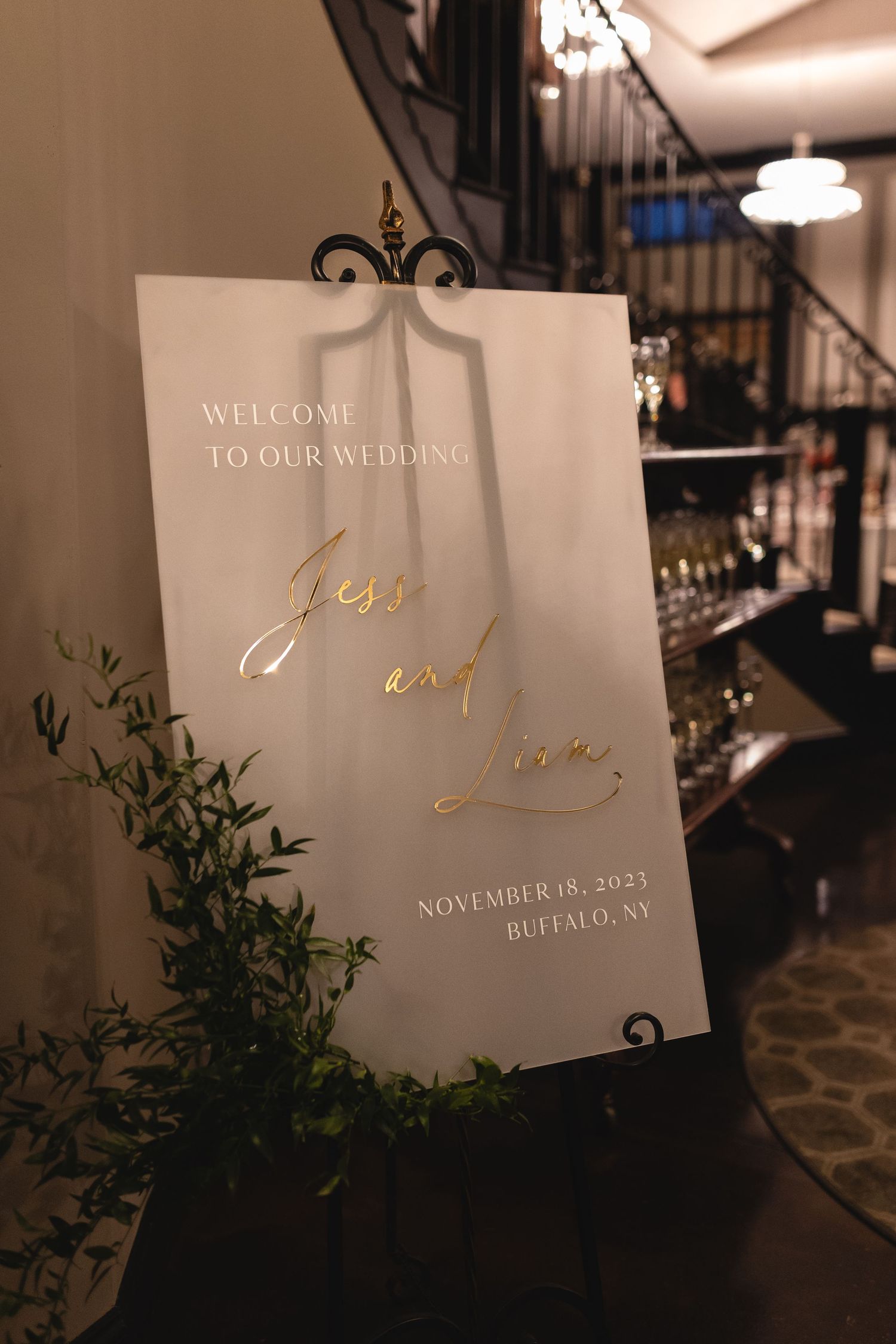 Elegant wedding welcome sign with gold text and greenery decoration displayed on a black ornate stand at indoor venue.