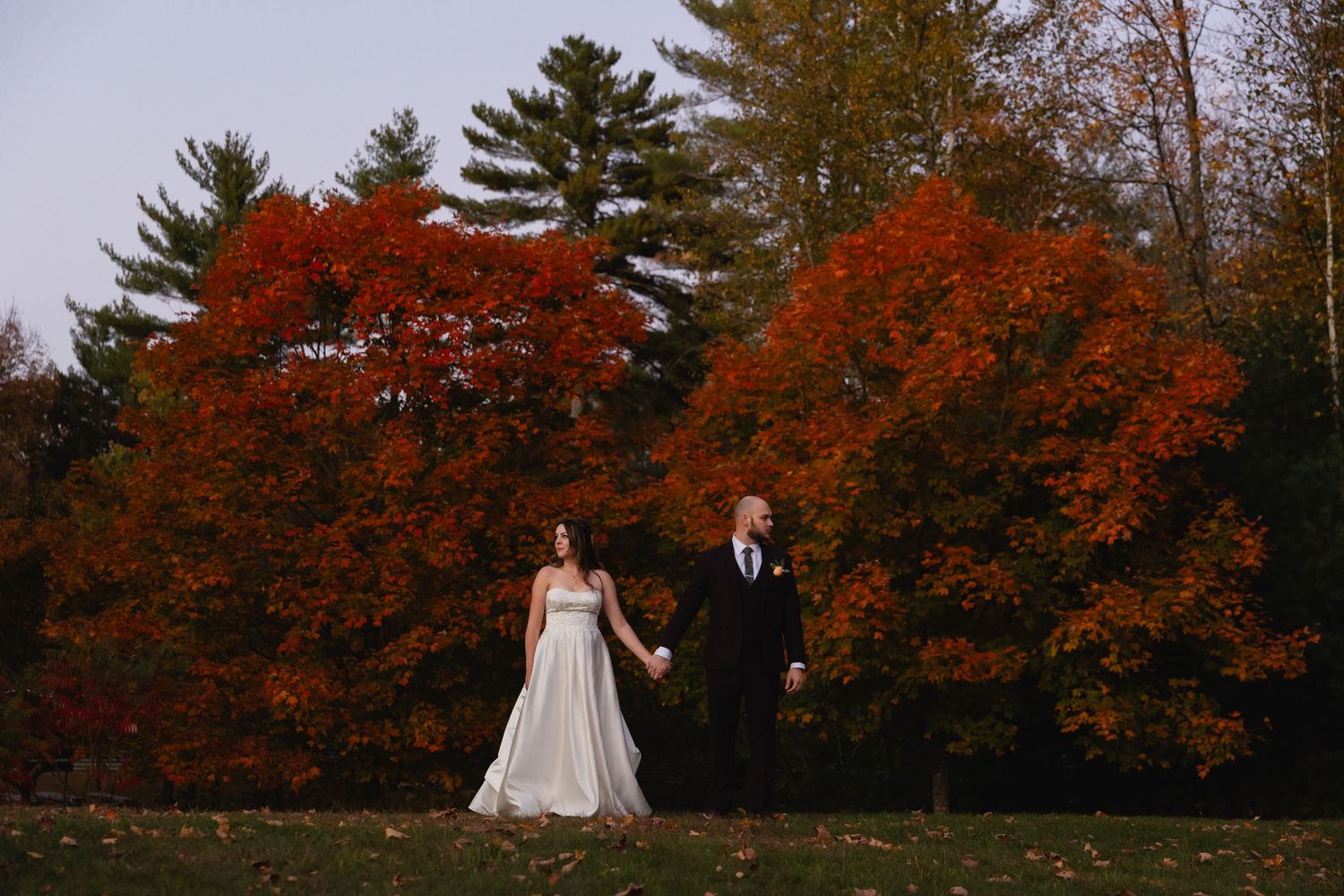 A couple in wedding attire stands together surrounded by vibrant red autumn foliage and evergreen trees.