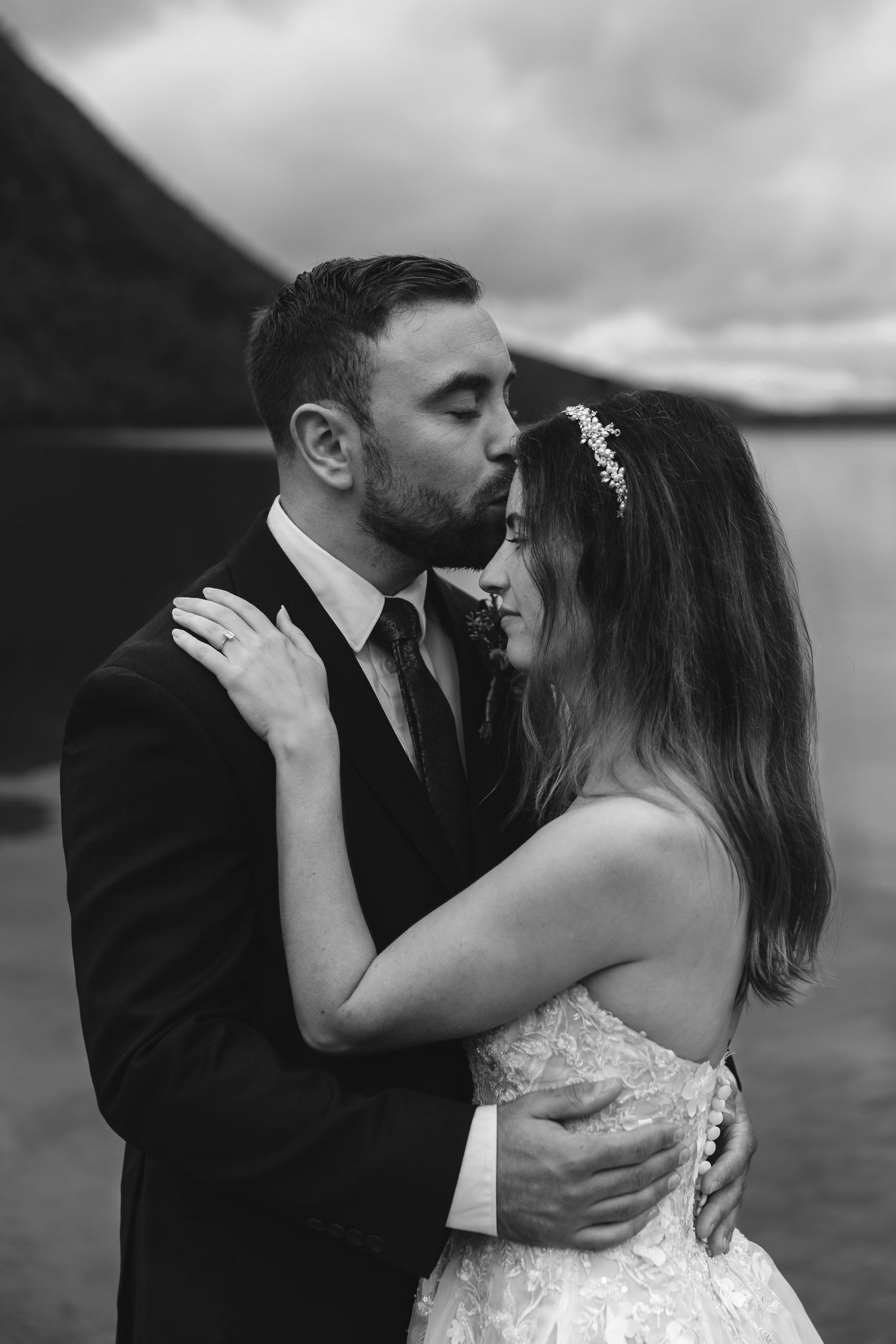 Romantic wedding couple embracing by a mountain lake on an overcast day.
