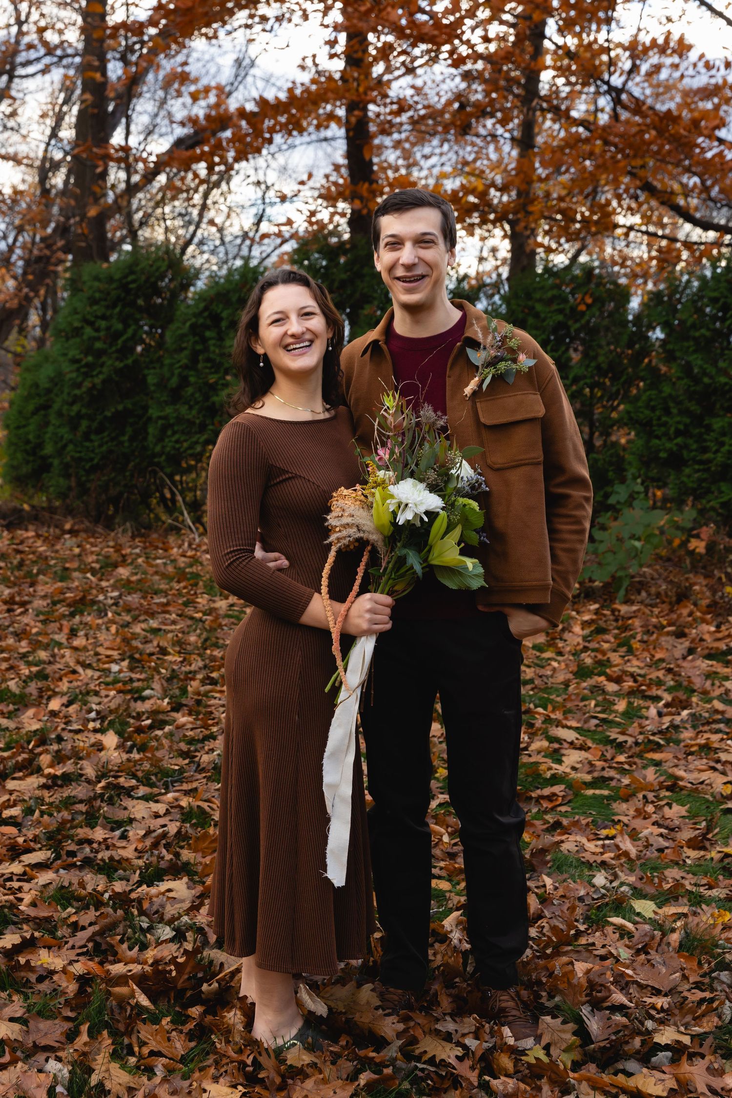 Two people stand together in a fall setting wearing brown clothing, holding a floral bouquet amid autumn leaves.