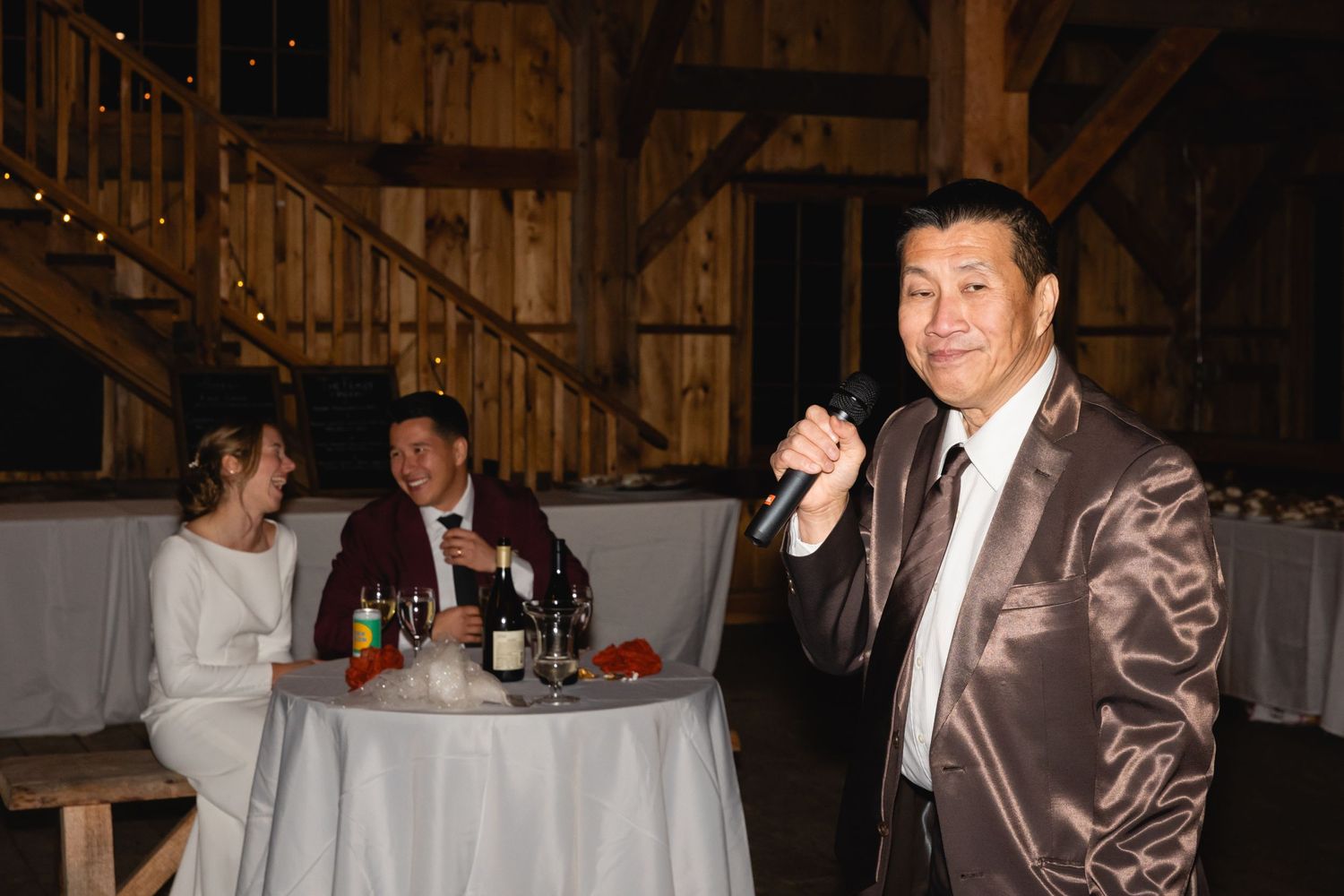 A person speaks into a microphone at a wedding reception while others sit at a decorated table in a rustic venue.