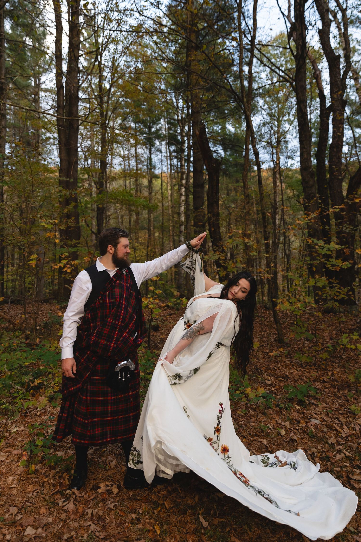 A couple in Scottish wedding attire strikes a playful dance pose in the woods.