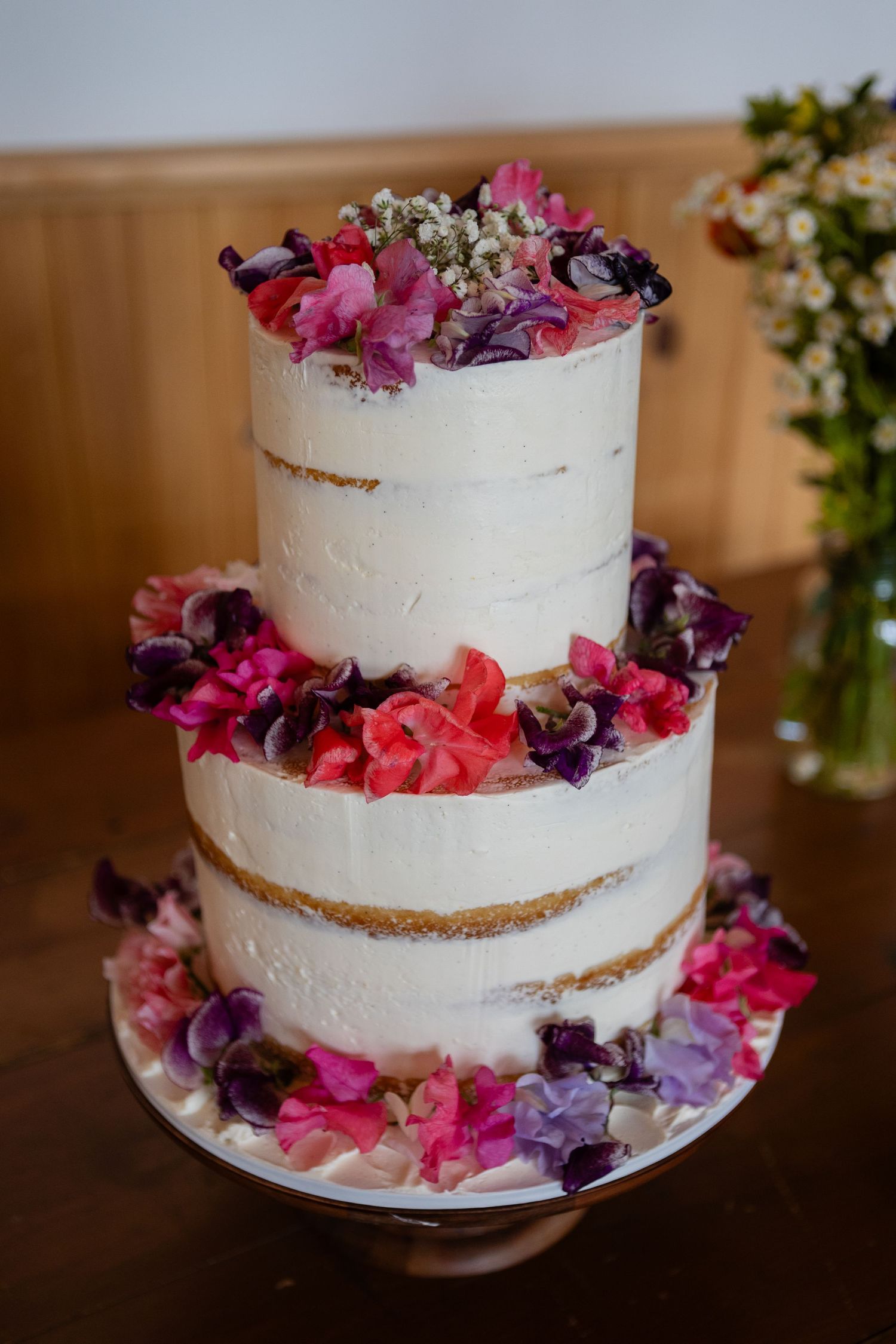A white semi-naked wedding cake decorated with pink and purple fresh flowers.