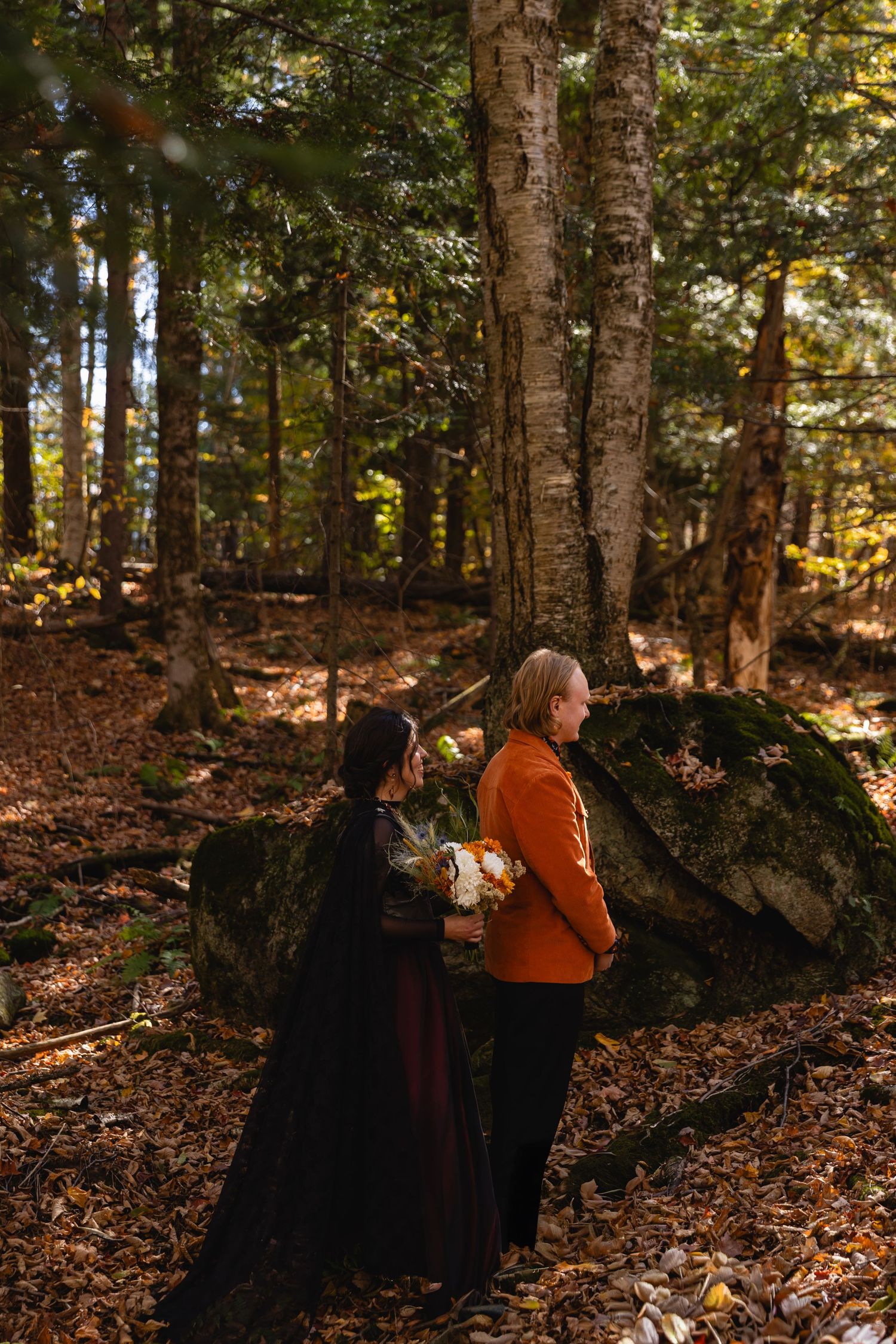 A couple shares a tender moment in a sunlit autumn forest with fallen leaves and tall trees surrounding them.