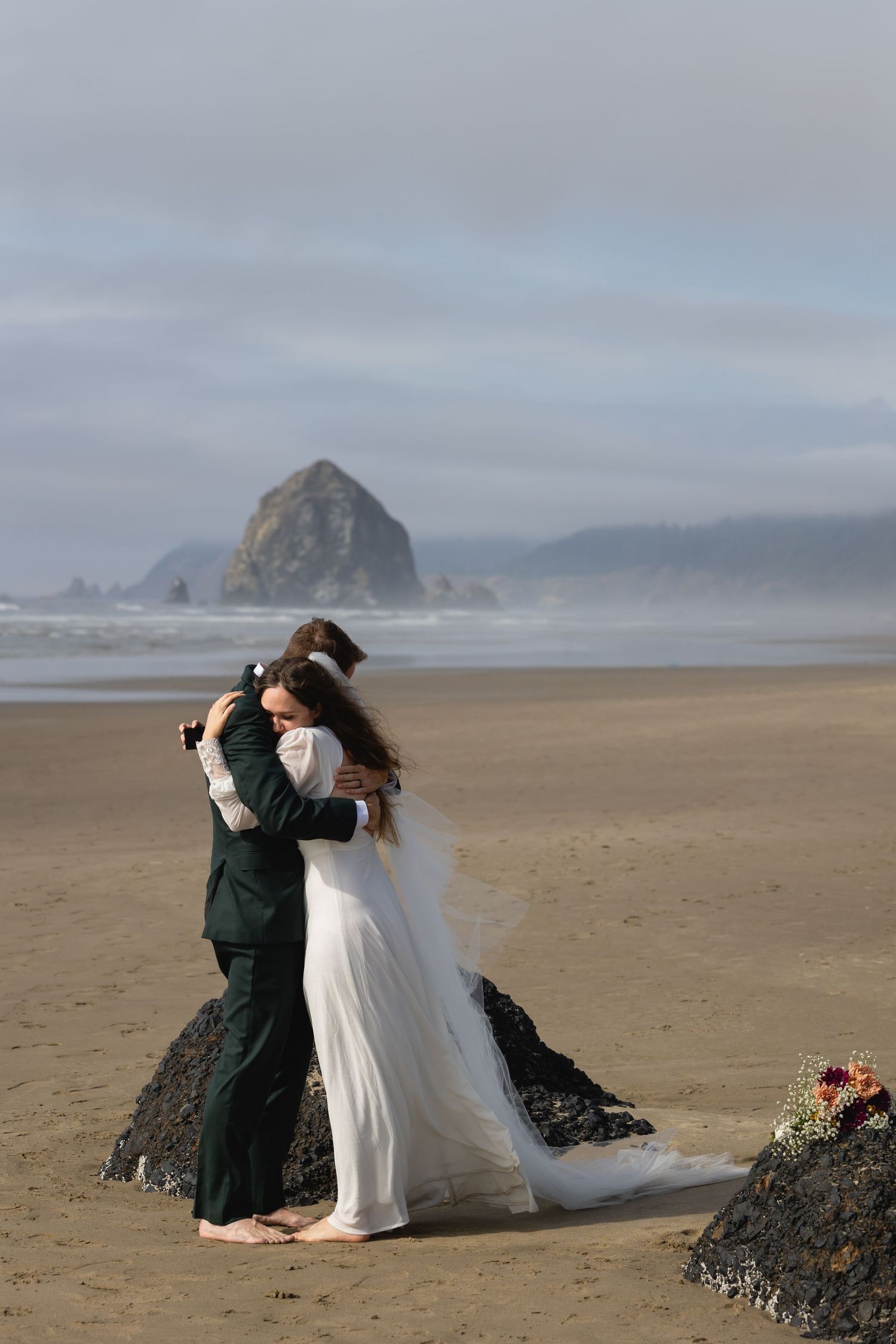 Two people in formal attire embrace on a misty beach with dramatic rock formations visible along the coastline.