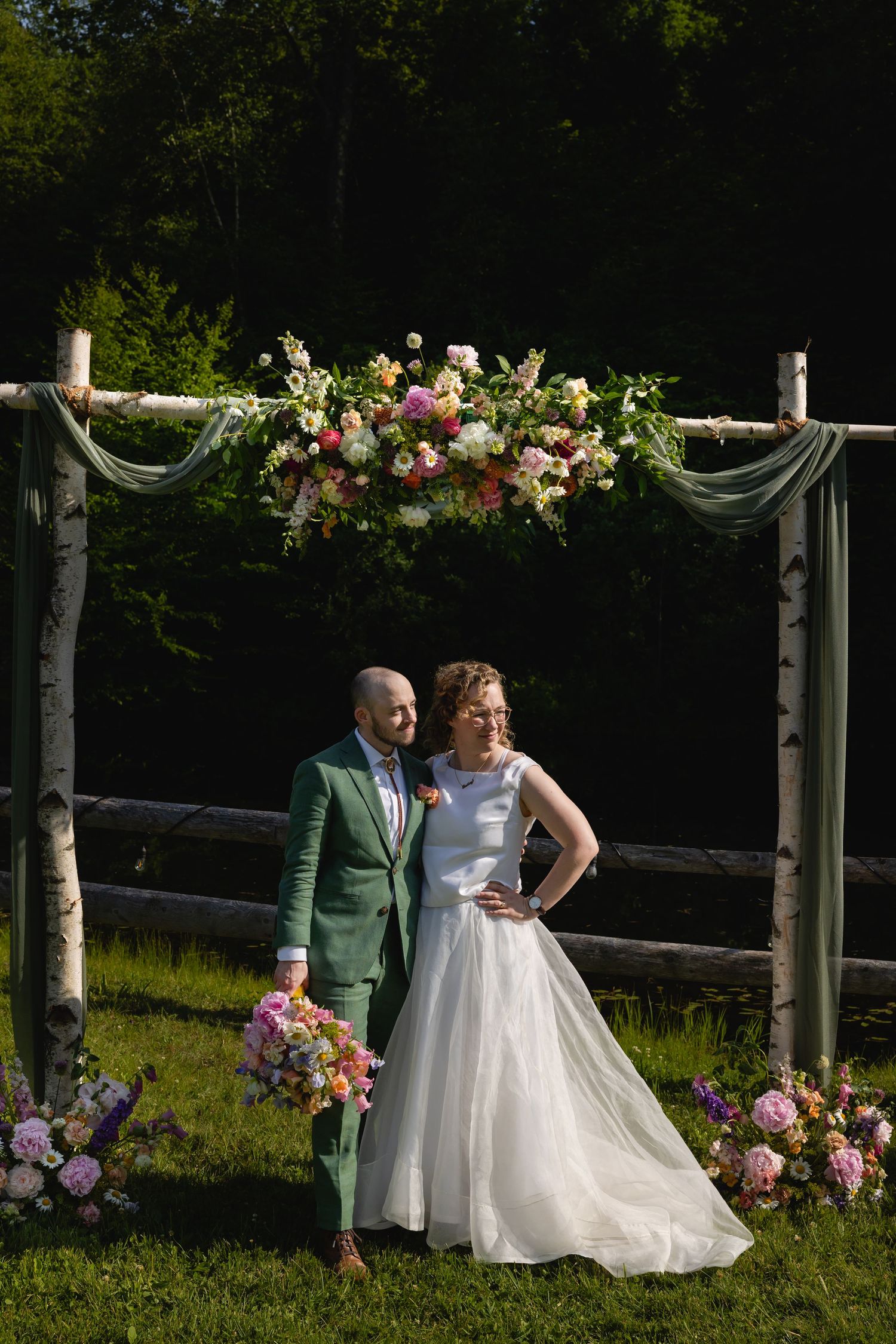 A couple poses under a floral wedding arch with pink roses and white fabric draping in a garden setting.