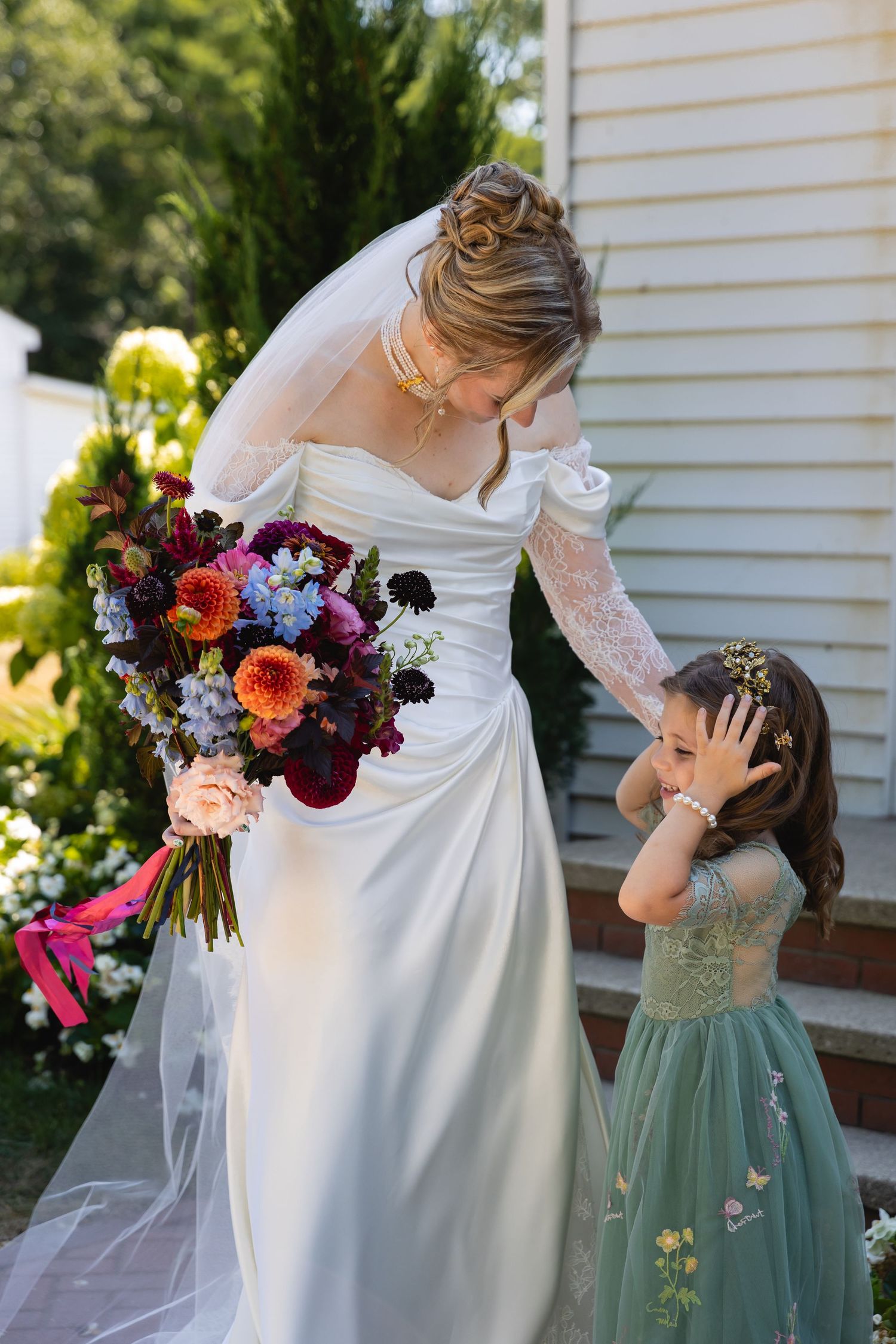 A bride in a white dress stands with a flower girl in sage green, holding vibrant autumn-colored bouquets.