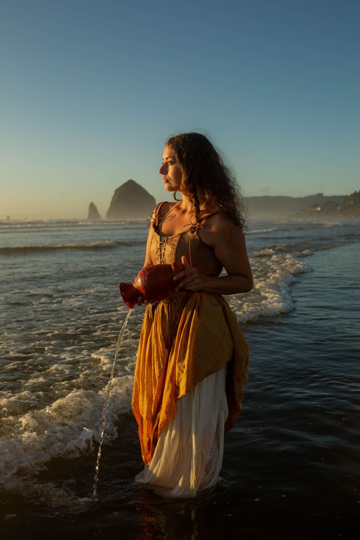 Woman in golden dress holding red vessel wading in ocean waves at golden sunset.