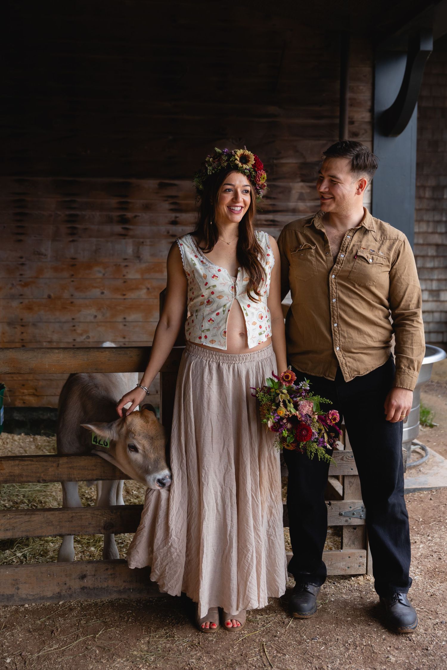 Smiling couple posing with a calf in a rustic barn, bride wearing floral crown and bouquet.