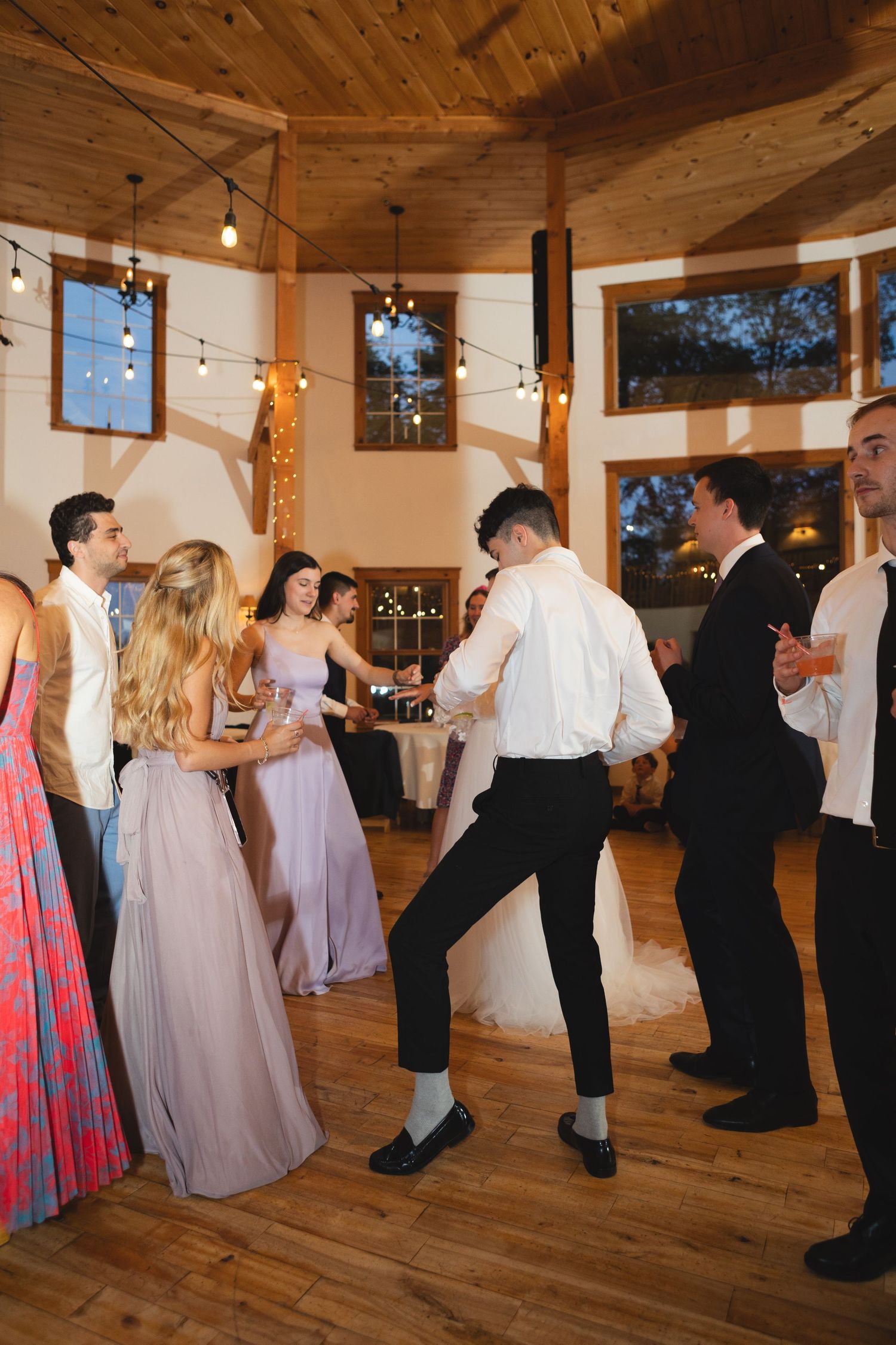 Wedding party dancing in formal attire under string lights in wooden interior venue.