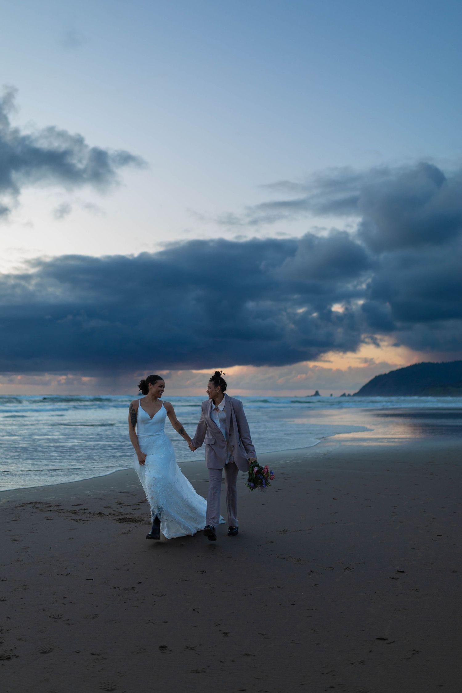 A couple strolls along a scenic beach at dusk holding a bouquet.