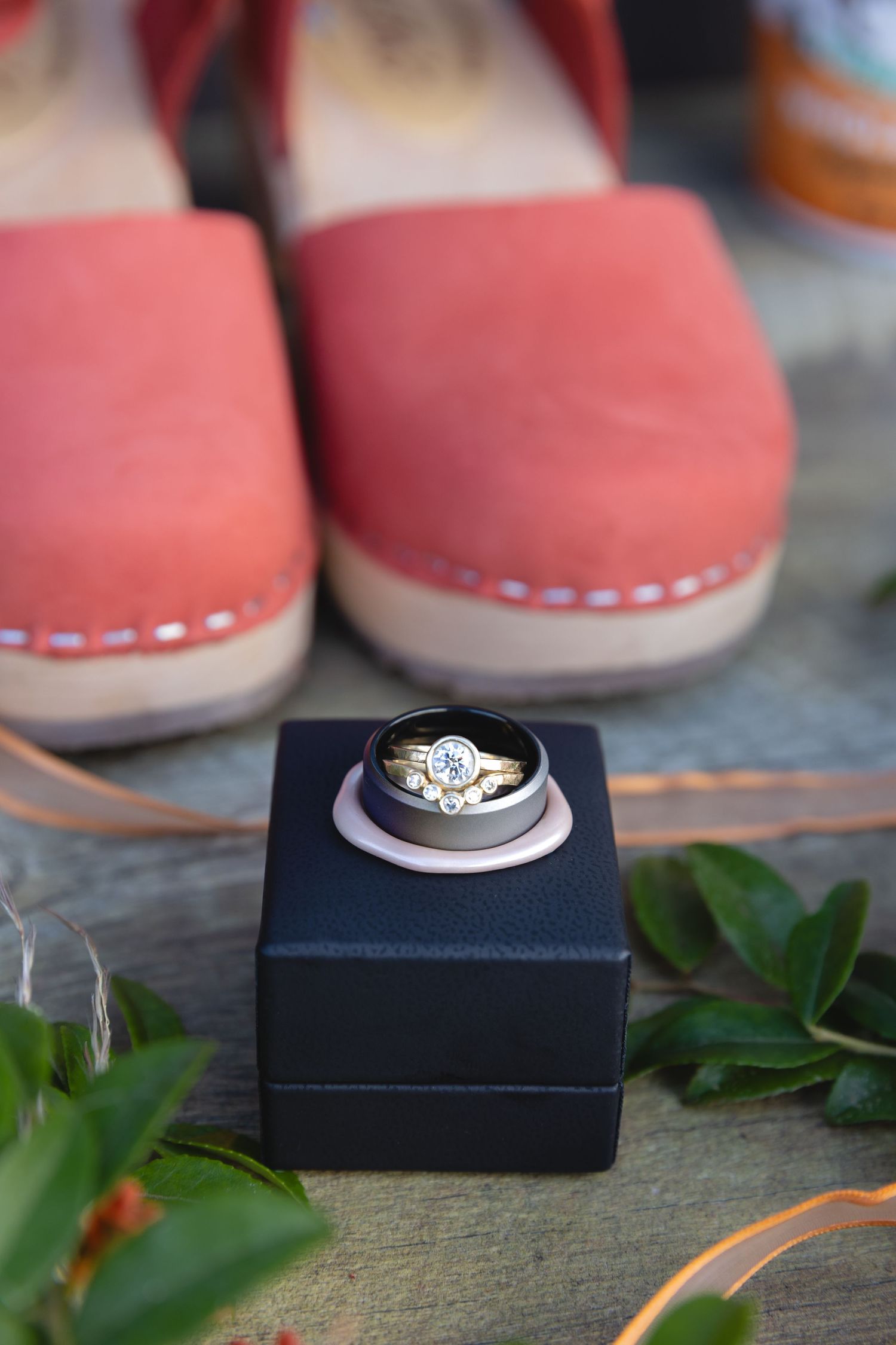 Diamond engagement ring displayed in navy box with coral shoes and greenery in background.
