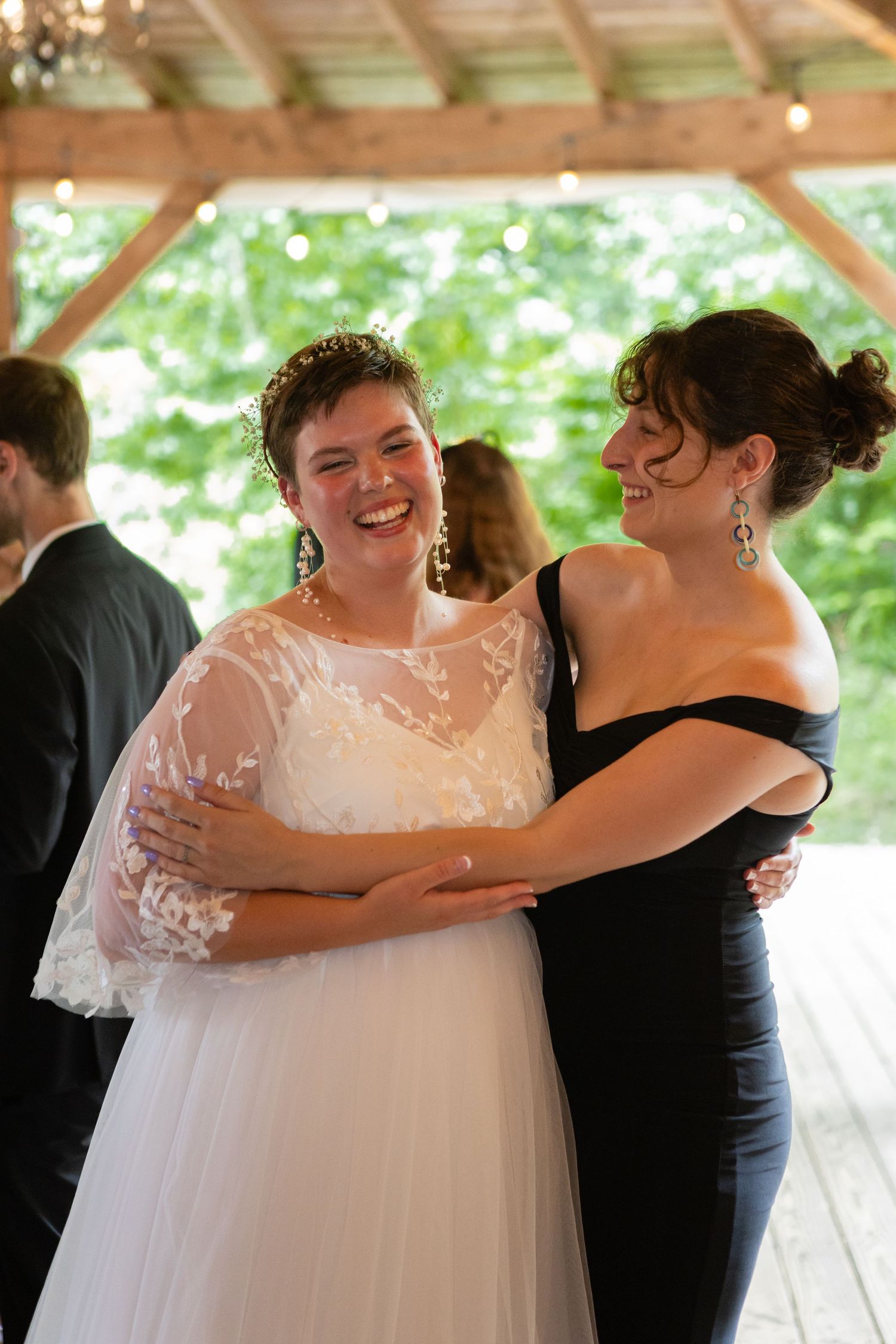 Joyful moment at wedding reception with bride and guest sharing laughter under wooden pavilion.