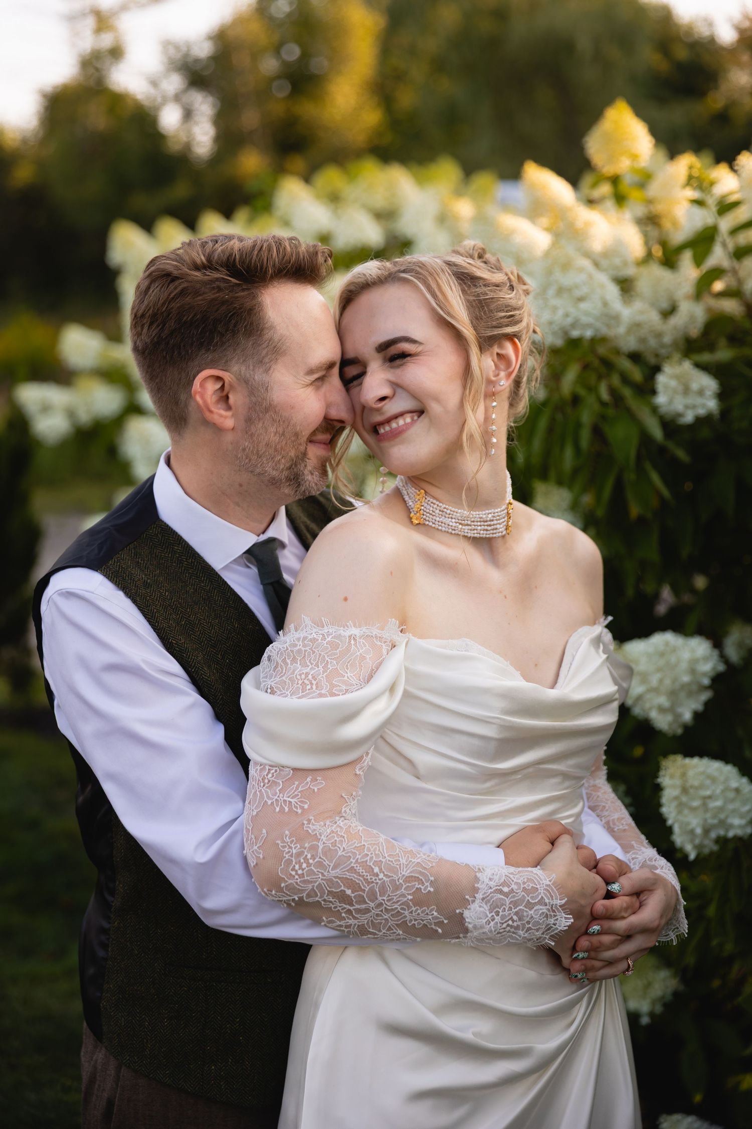 A couple shares intimate moments in their wedding attire against a backdrop of white flowers at sunset.