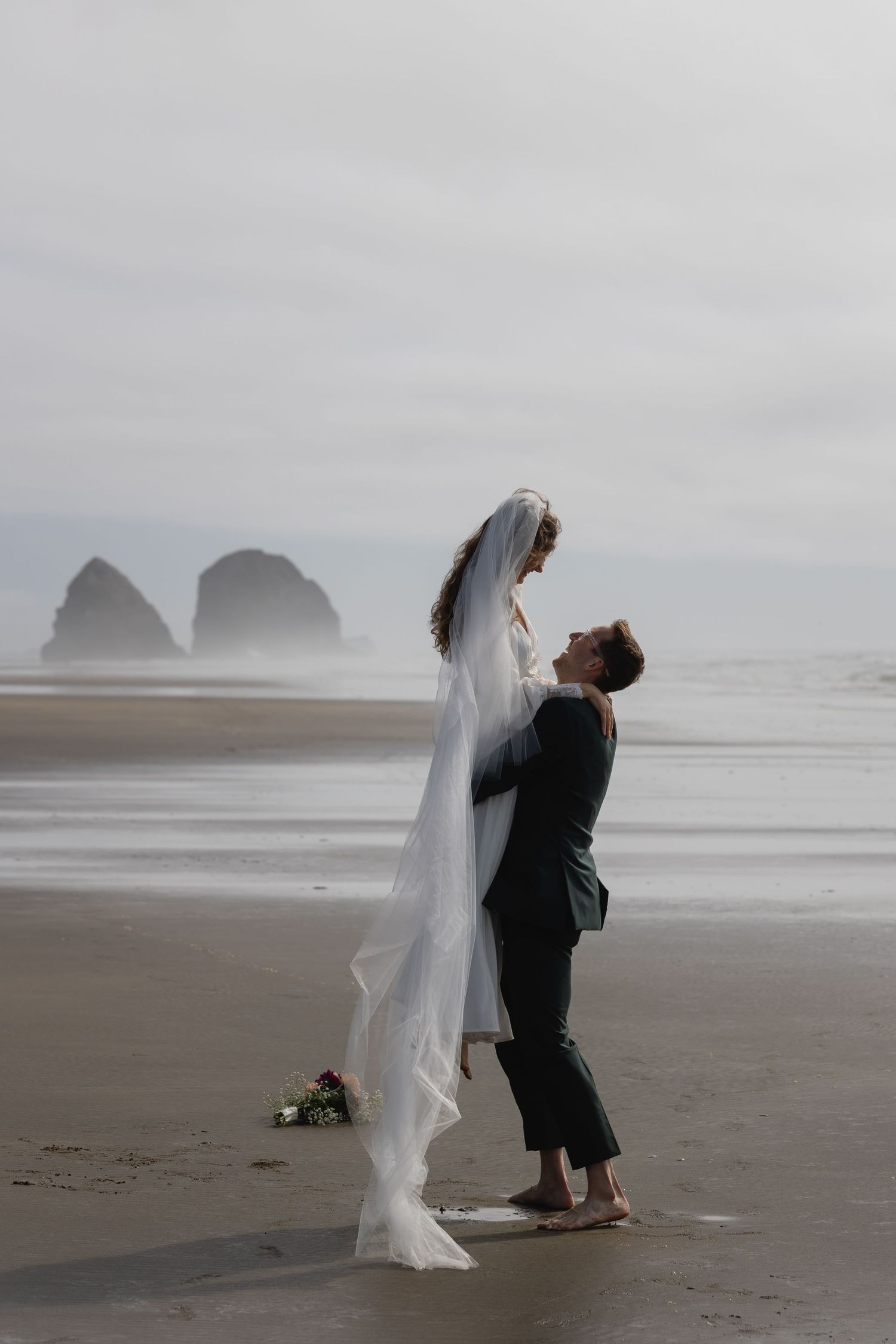 A romantic wedding couple embraces on a misty beach with dramatic rock formations in the background at Cannon Beach, Oregon.