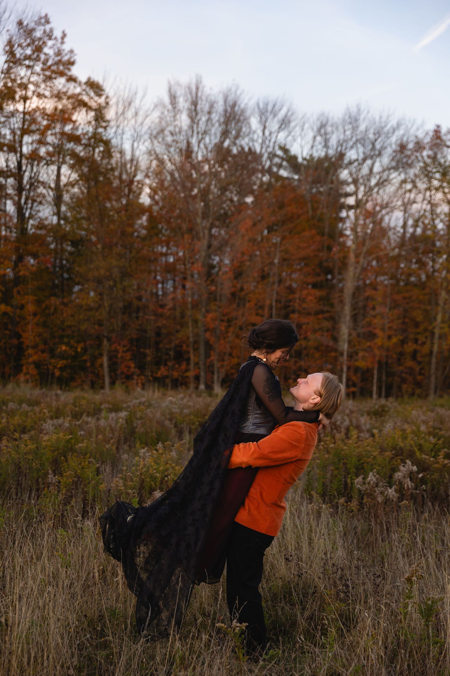 Two people in autumn attire dance together in a field with fall foliage and bare trees in the background.