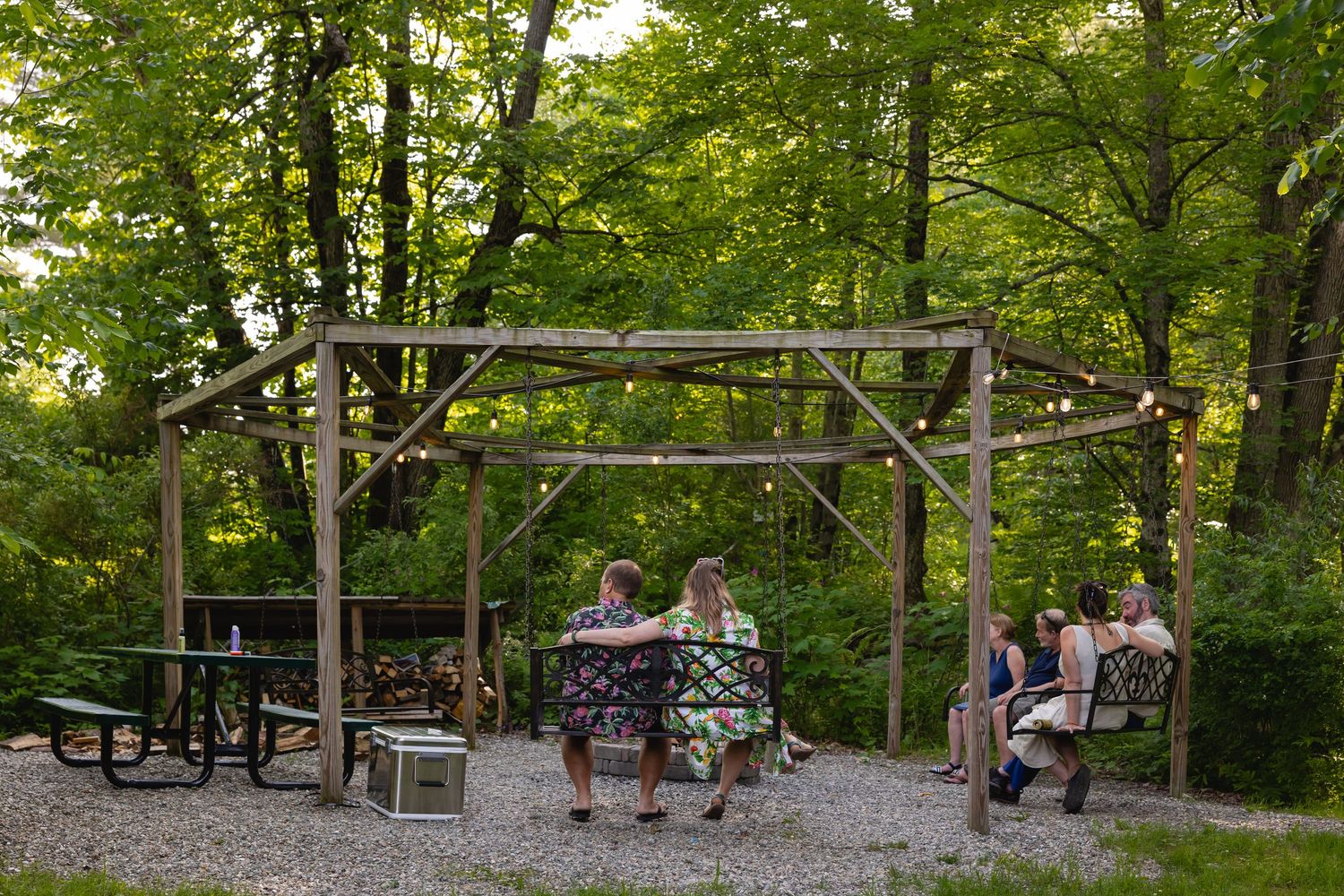 People relax at a rustic wooden pavilion with benches surrounded by lush green forest in a peaceful outdoor setting.