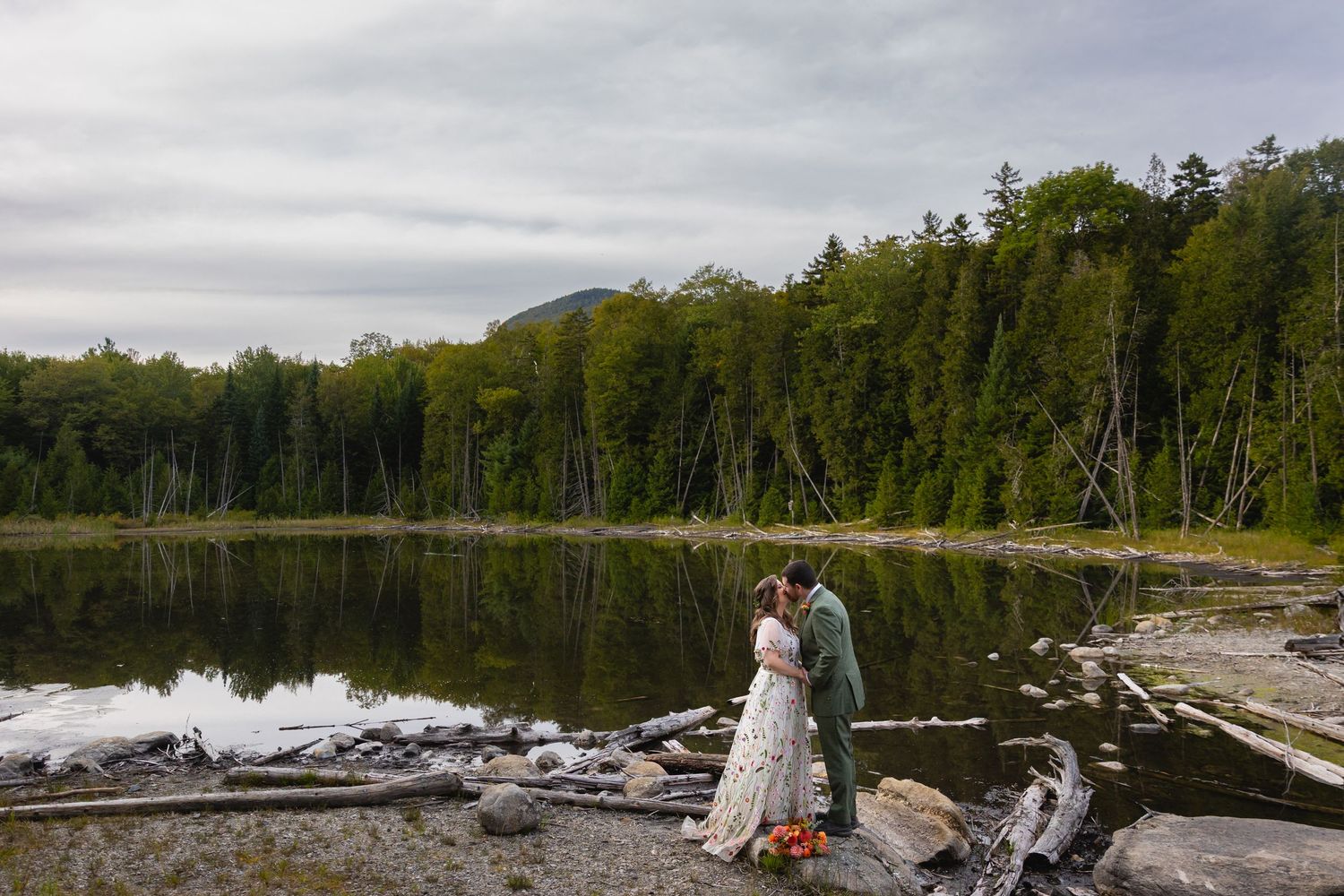 A scenic wedding photo shoot on a rocky lakeshore with mountains and forest in the background.