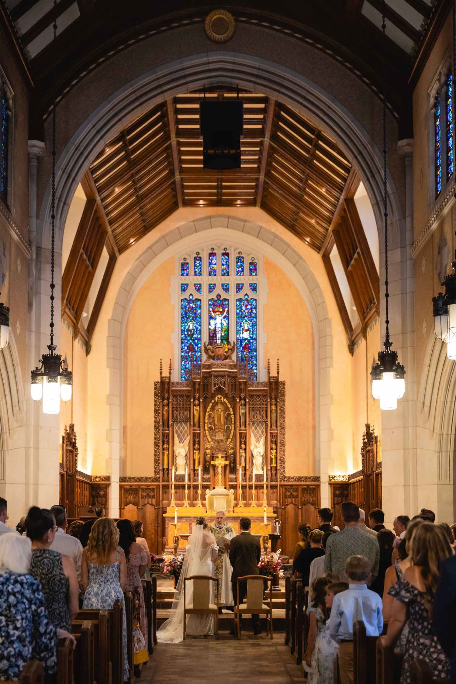 A church ceremony takes place under a vaulted ceiling with stained glass windows and wooden beams in an ornate sanctuary.