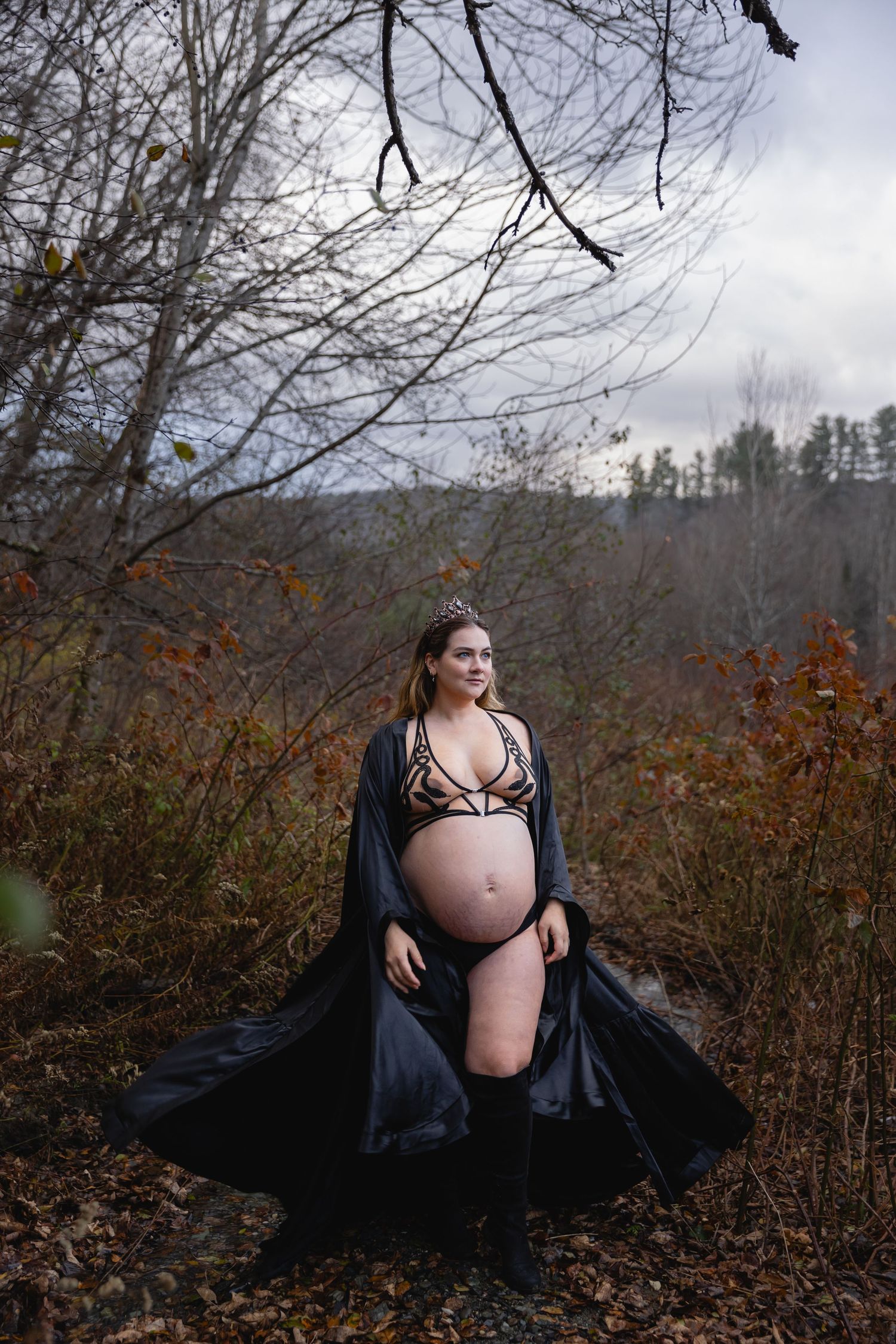 Pregnant woman in black lingerie and flowing cape poses outdoors in autumn forest with mountain backdrop.