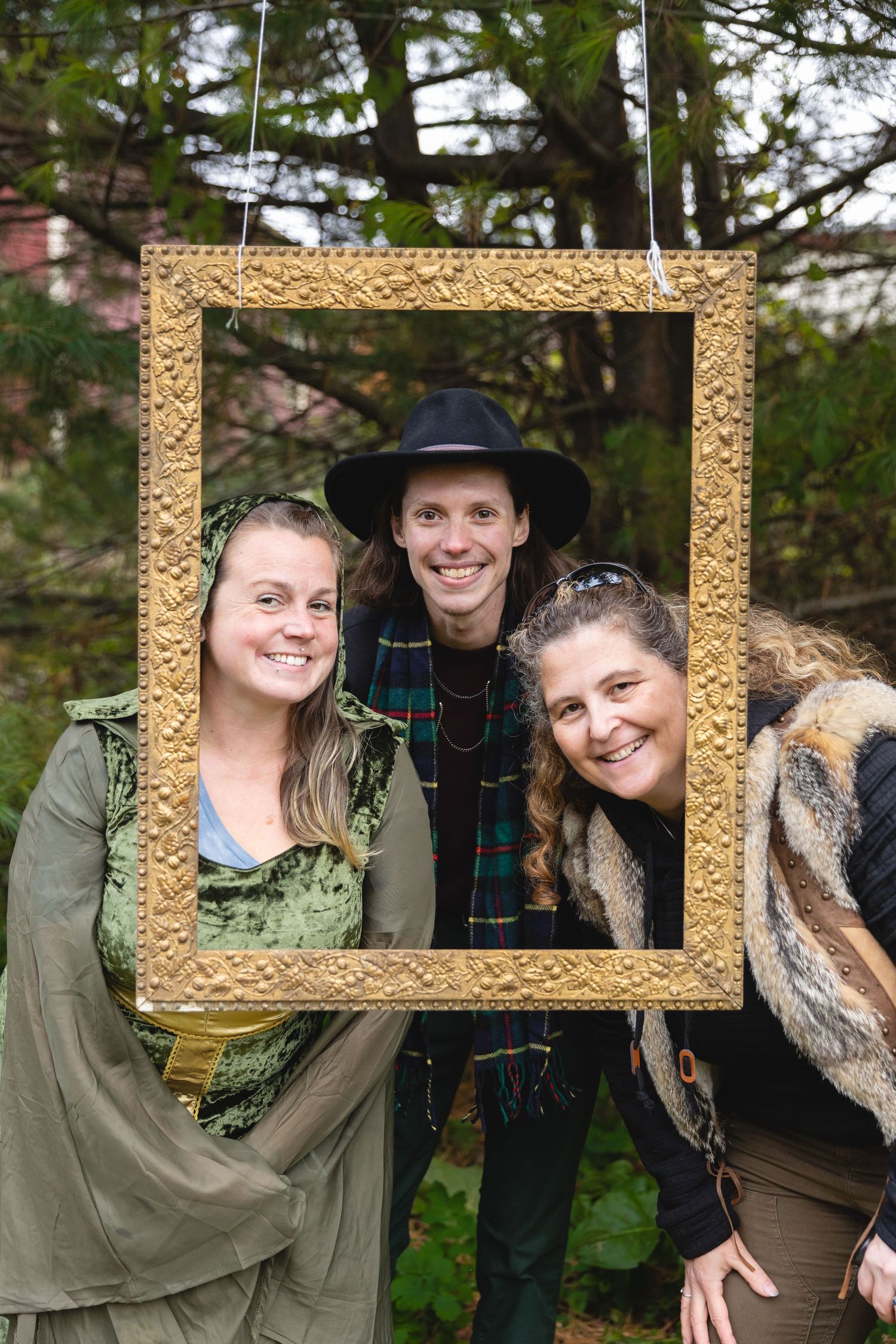 Three people posing playfully through decorative gold picture frame outdoors.