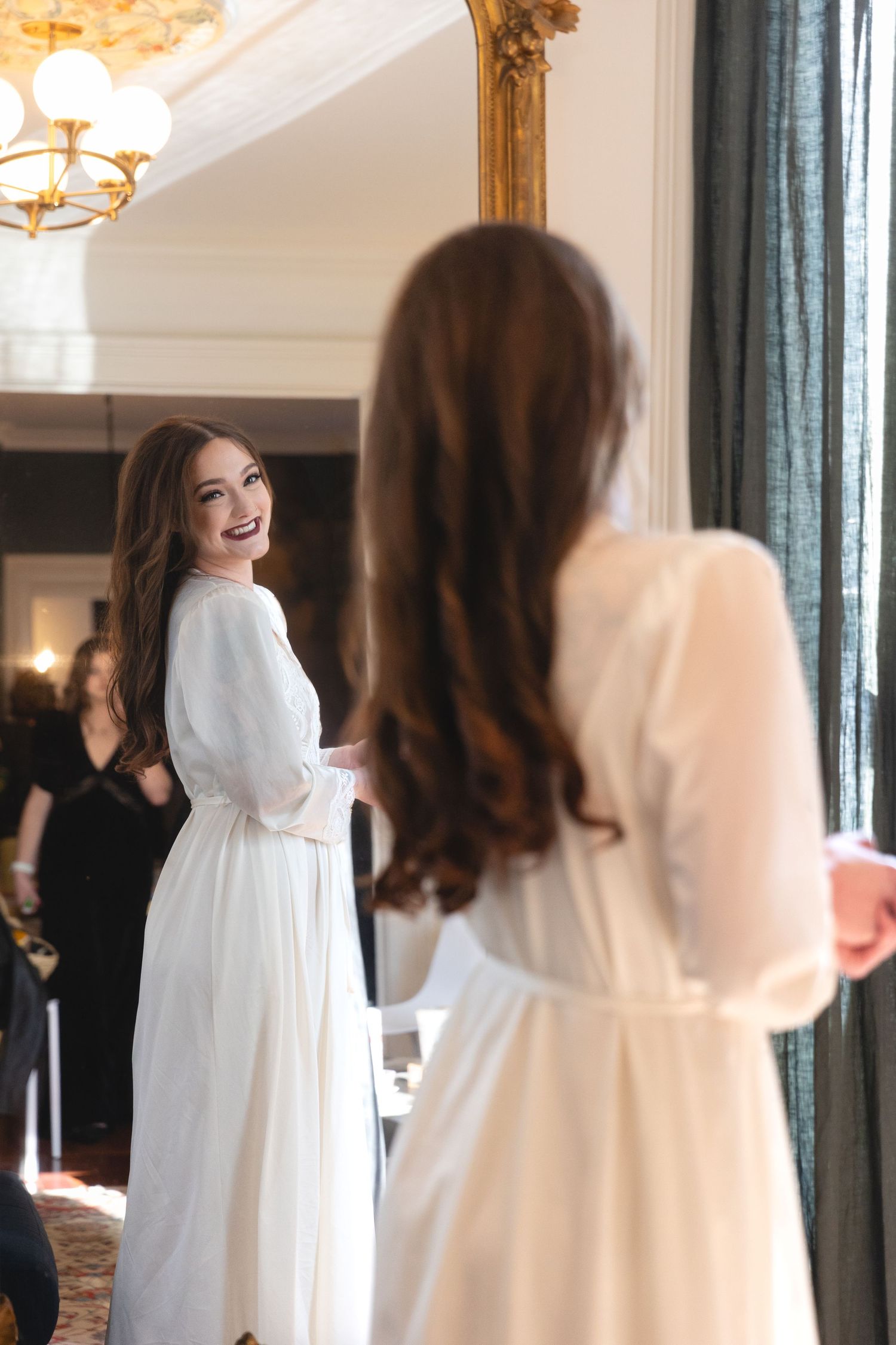 A person in a white dress stands near a mirror in an elegant room with an ornate gold frame and chandelier.