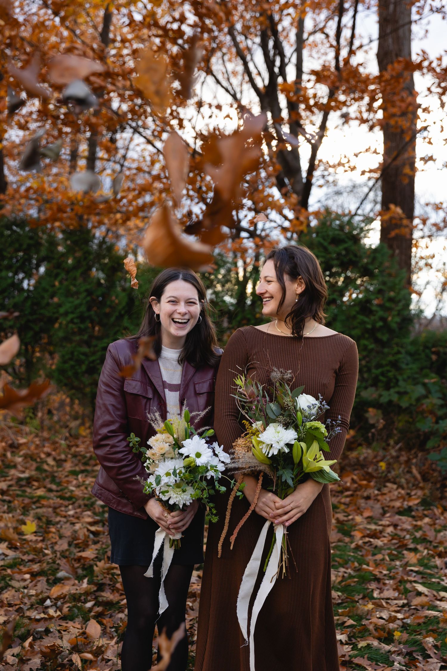 Two people stand laughing together holding white floral bouquets amid fallen autumn leaves in a woodland setting.