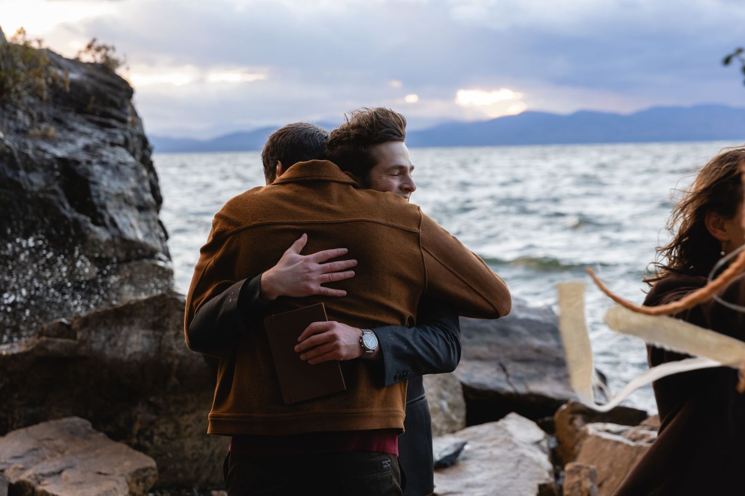 Two people embrace in a warm hug near rocky cliffs by the ocean with moody clouds and mountains in the distance.