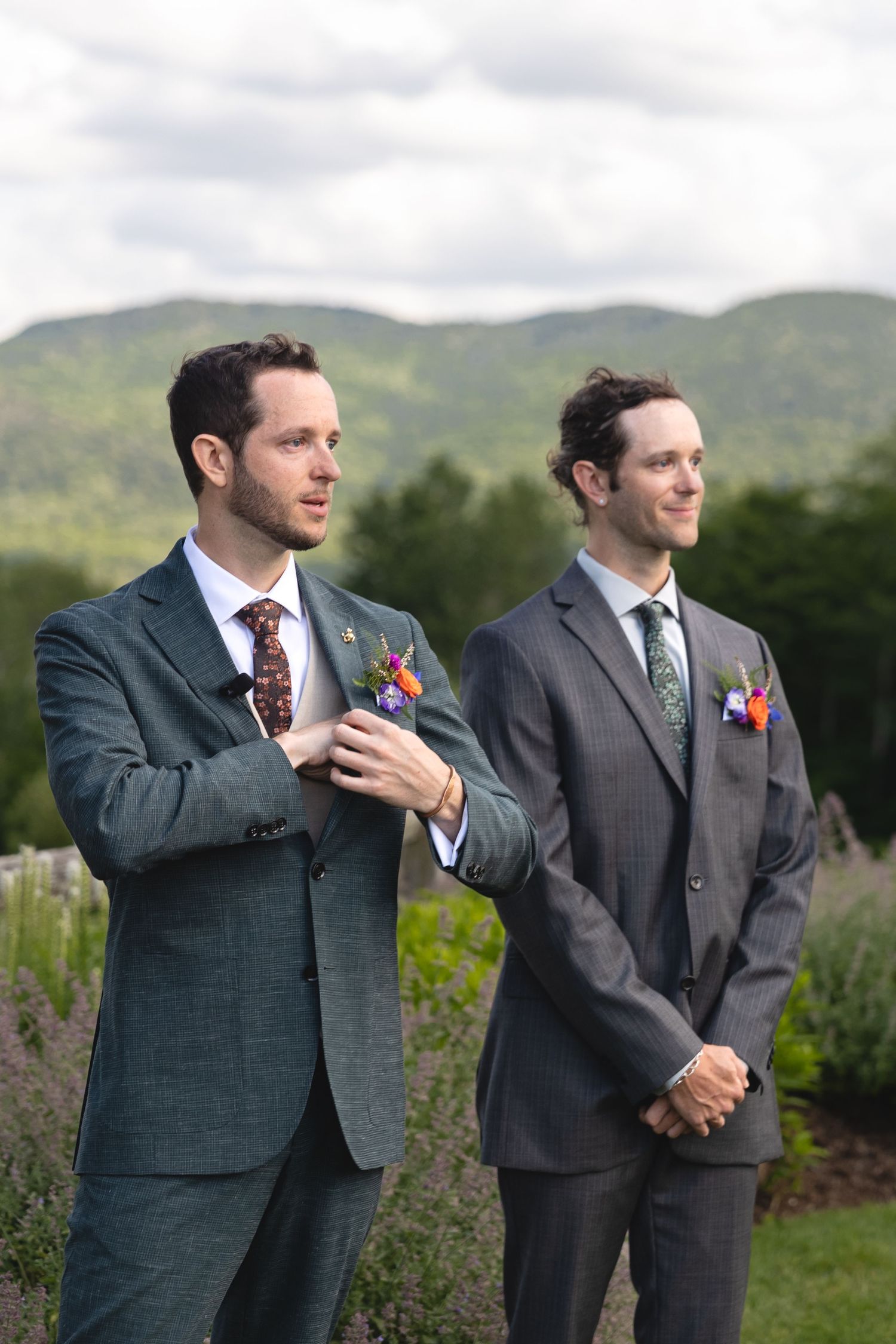 Two men in formal suits stand together during an outdoor wedding ceremony with mountains in the background.