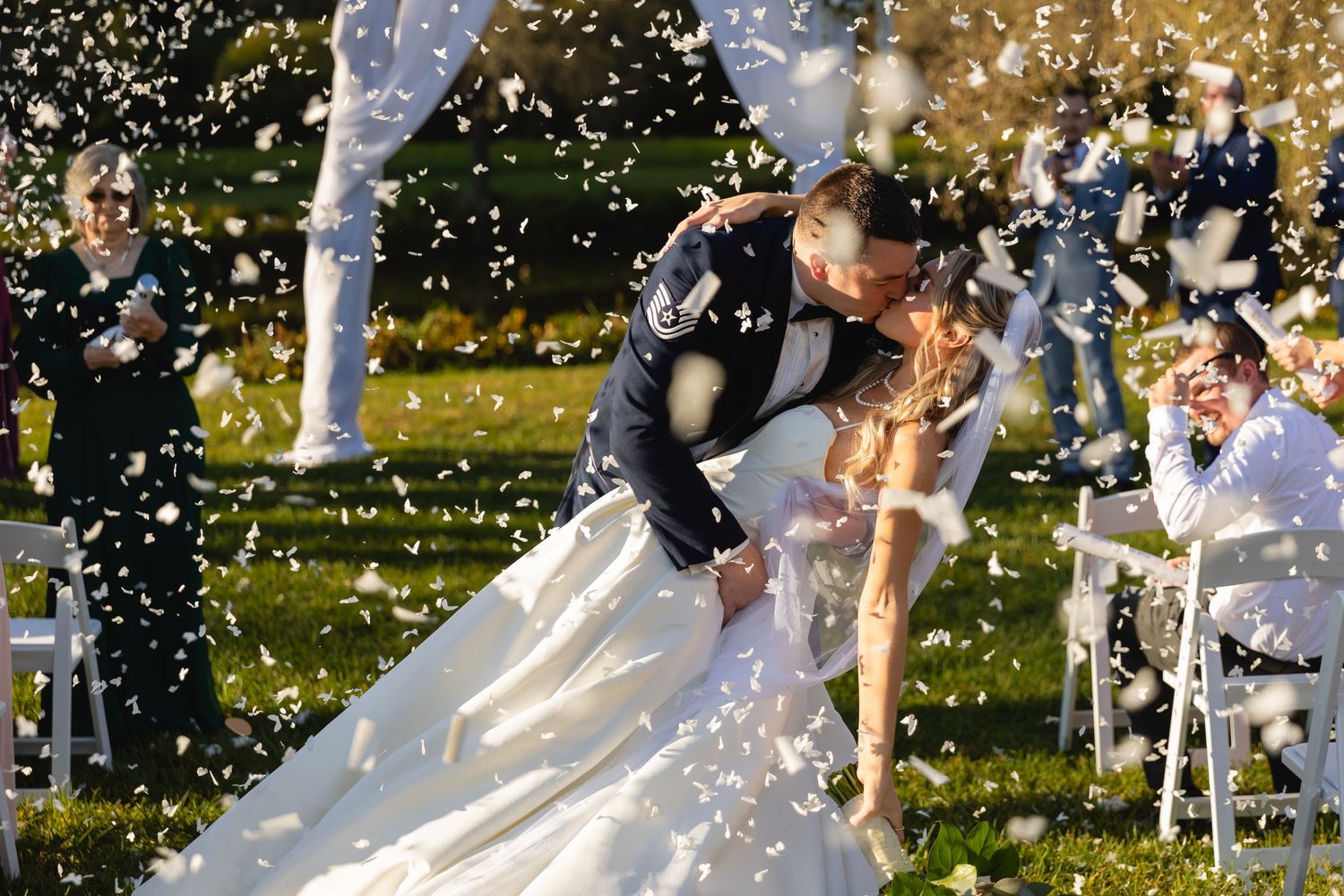 A couple shares their first dance as newlyweds while white flower petals shower down around them on a sunny day.