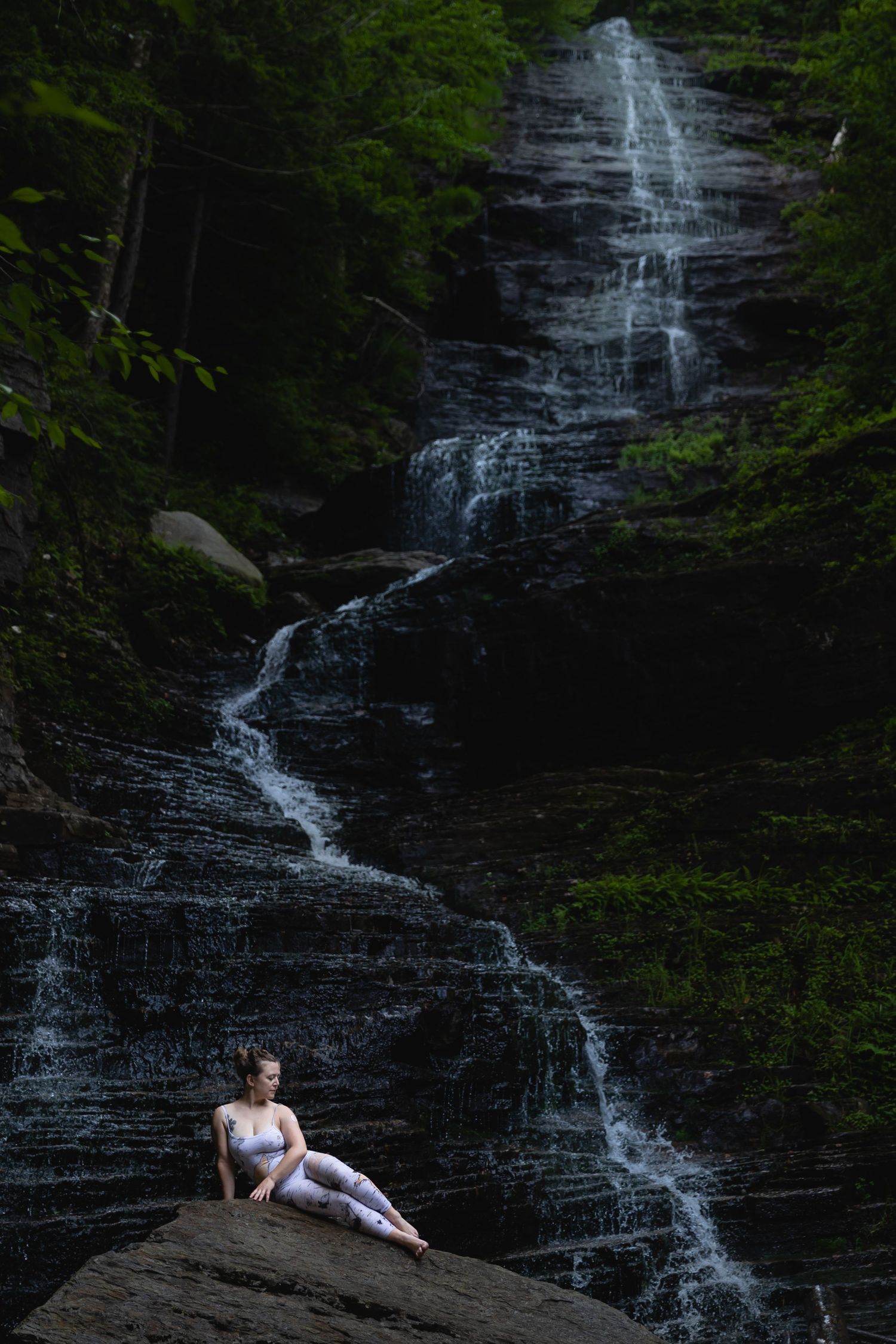 Woman in athletic wear sits on rock before cascading forest waterfall.
