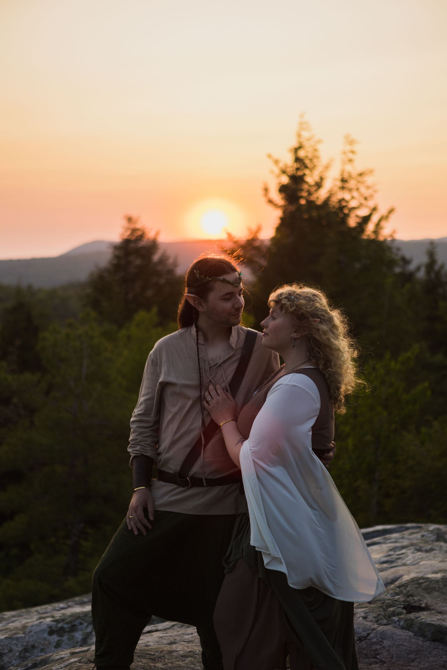 Couple in fantasy costumes sharing romantic moment on rocky hilltop at golden sunset.