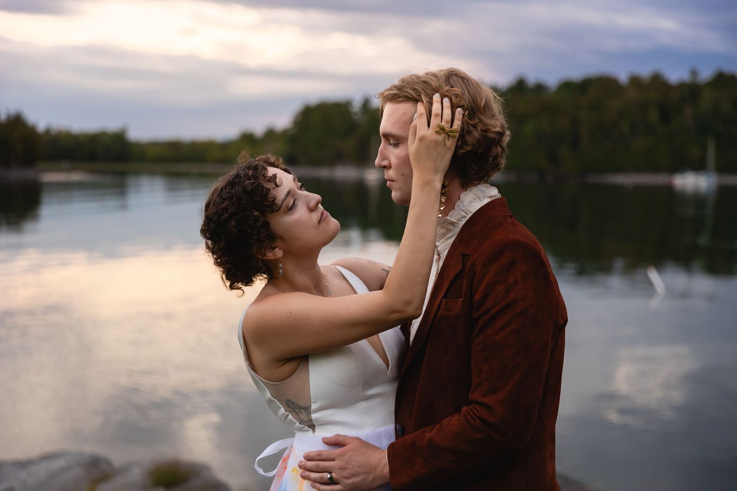 Couple embracing intimately by a lake at dusk, woman gently touching mans hair in soft light.