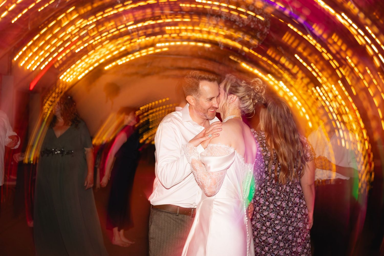 A couple dances together at a wedding reception with colorful lighting and other guests in the background.