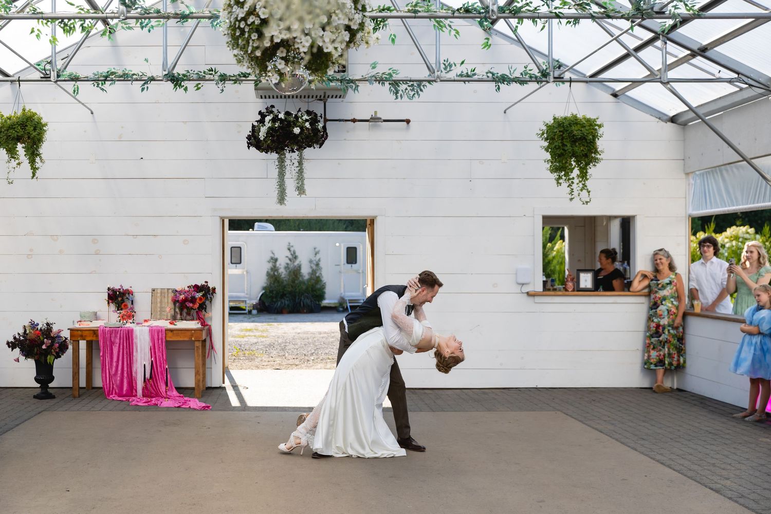 A couple performs a romantic dance dip on a covered outdoor patio with hanging plants and guests watching nearby.