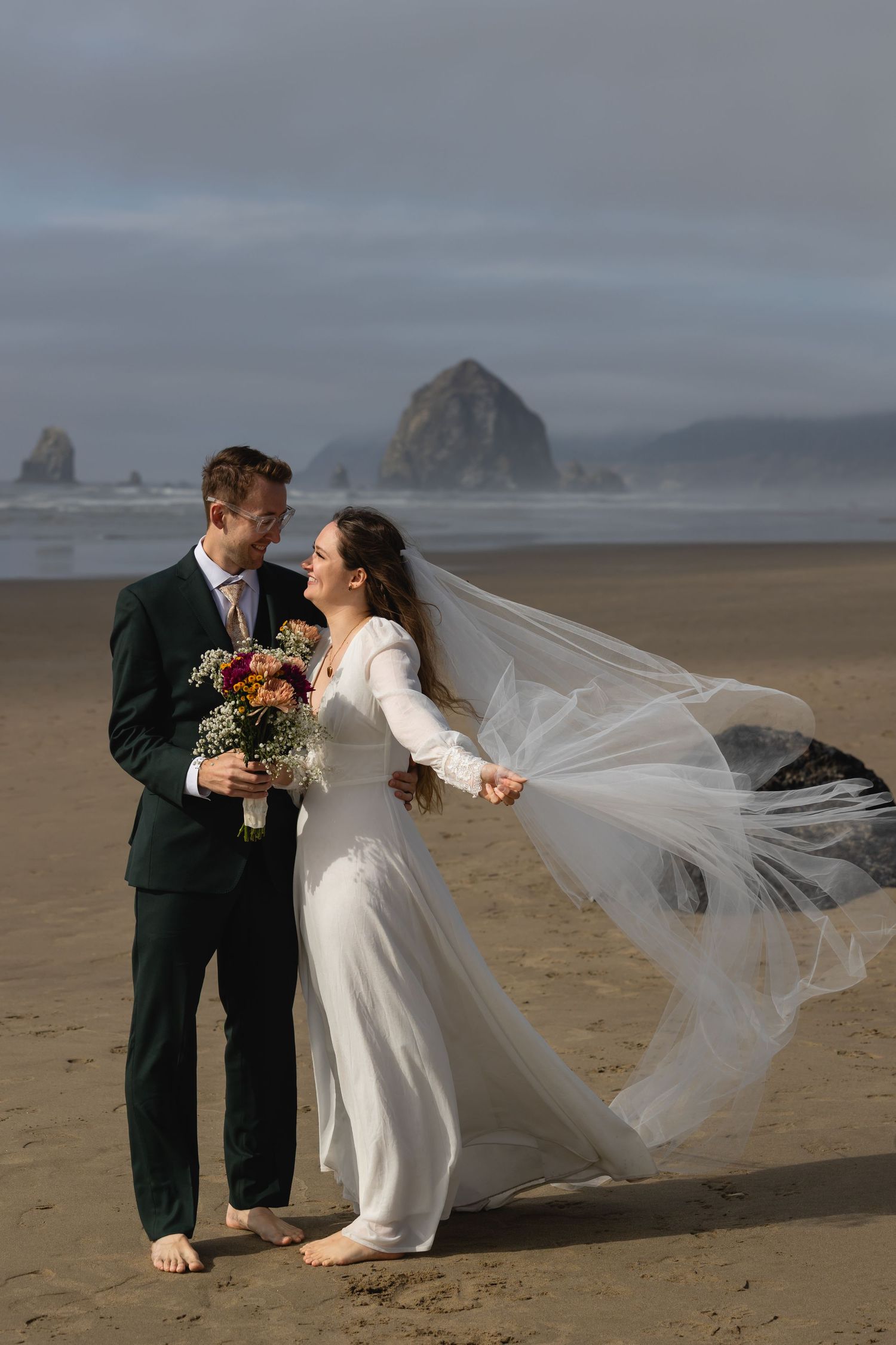 A couple in wedding attire shares a romantic moment on a misty beach with dramatic coastal rock formations in the background.