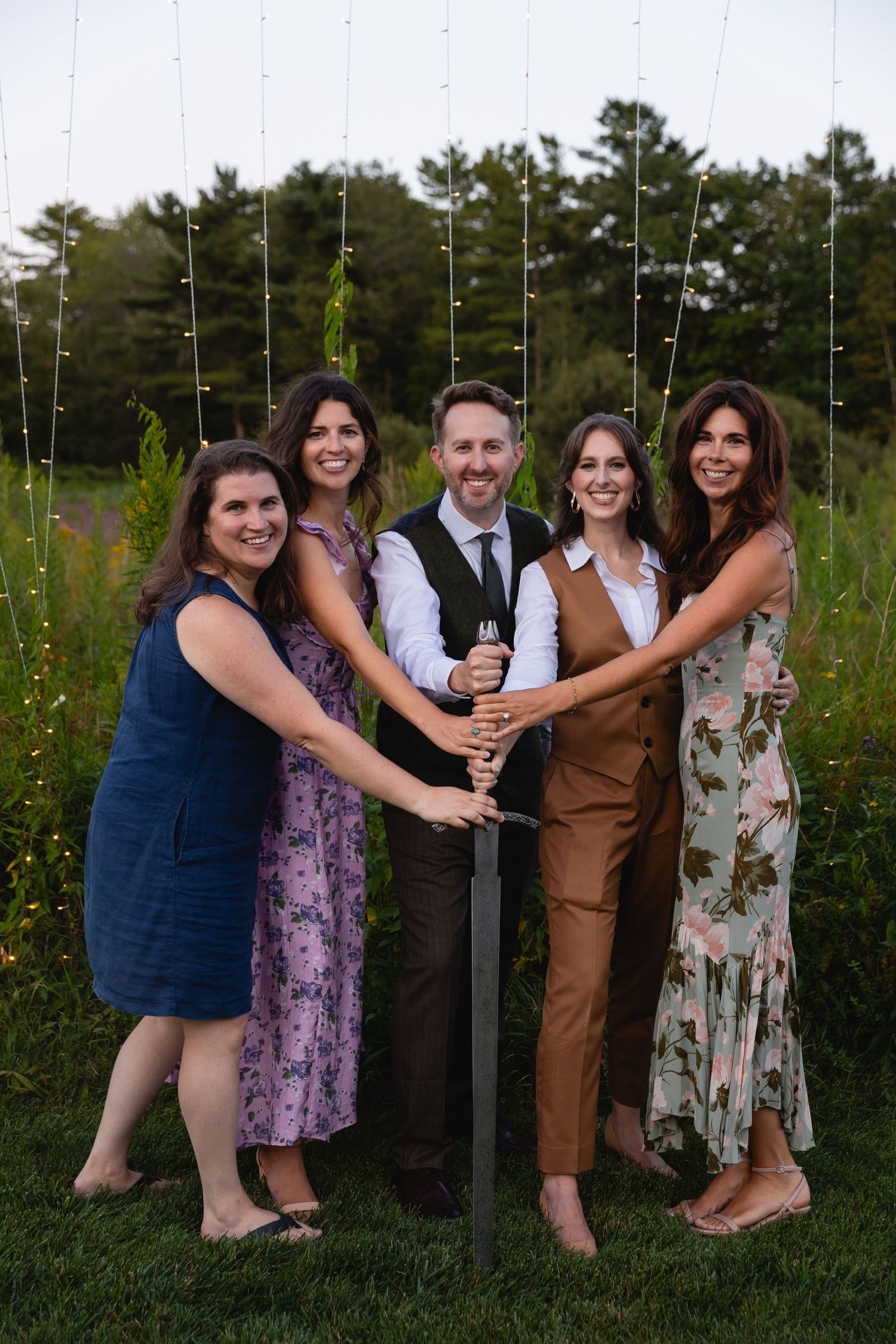 A group poses together outdoors in formal attire against a backdrop of trees and string lights at dusk.