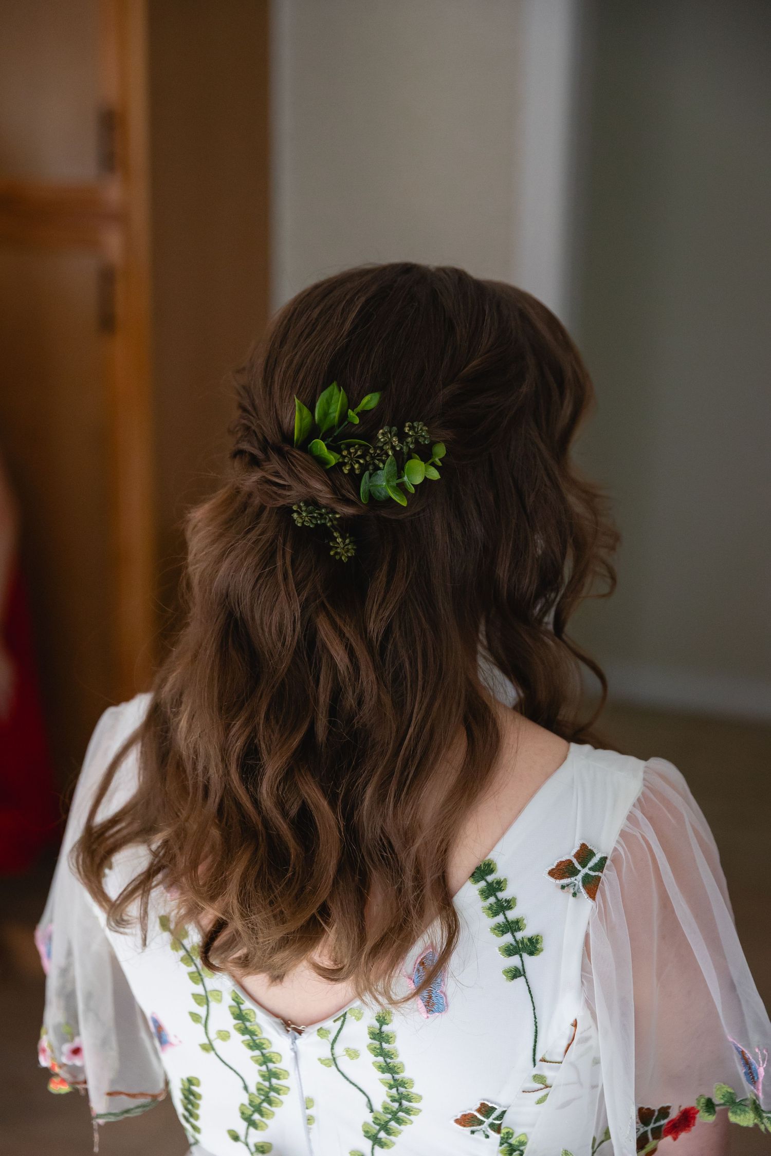 Person wearing a white dress with floral embroidery and green leaves in their wavy brown hair.