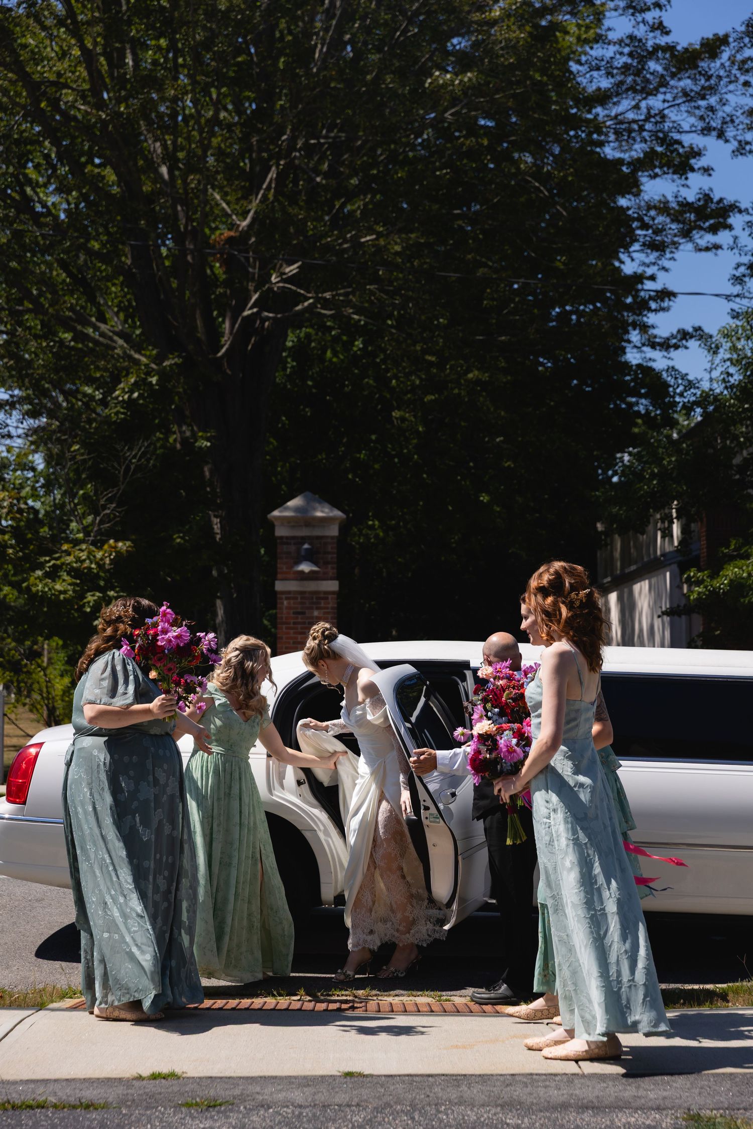 A group in formal attire gathers outside near parked cars on a sunny day, creating a candid moment.