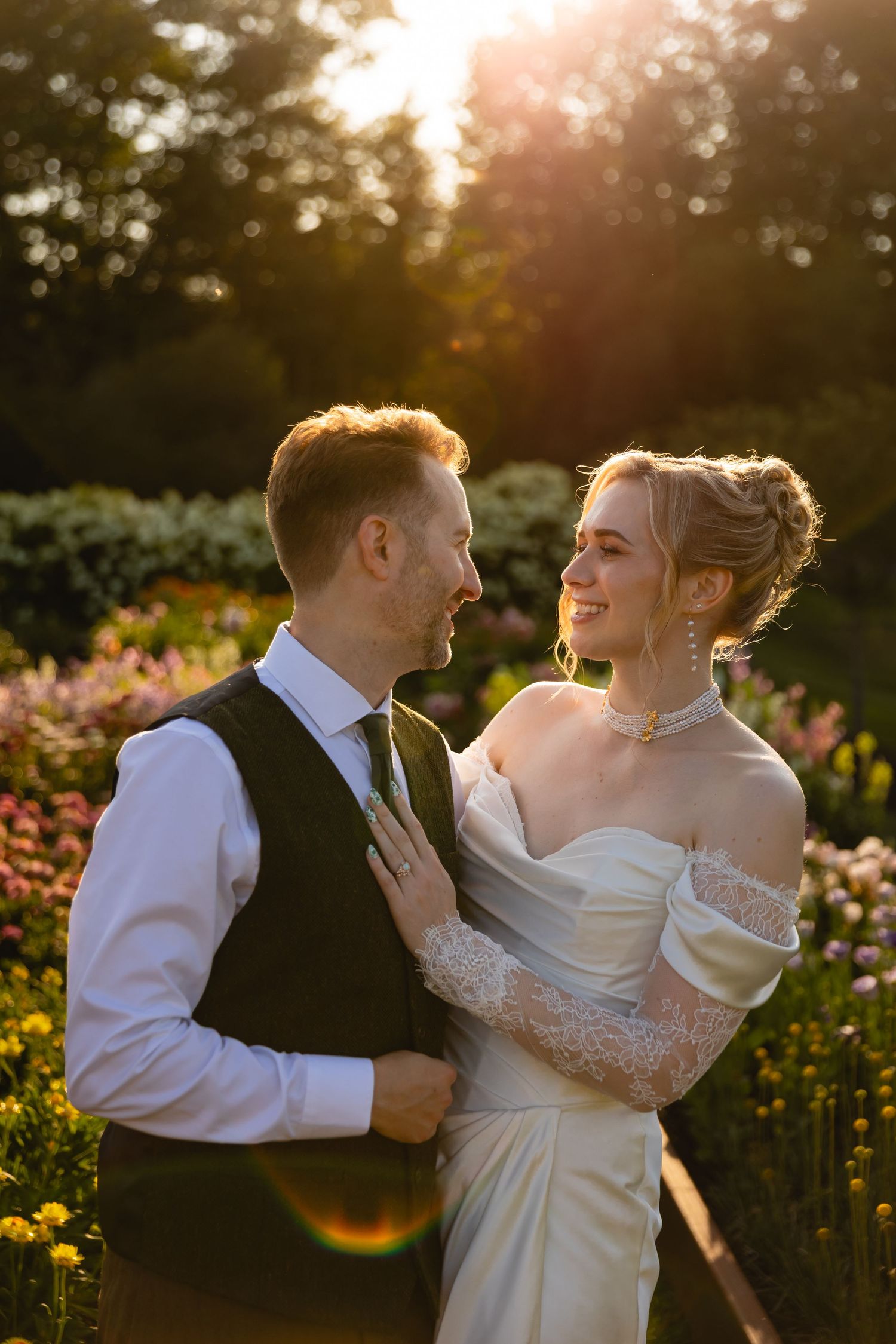 A romantic wedding couple embraces in a garden at sunset with golden sunlight streaming through the trees.